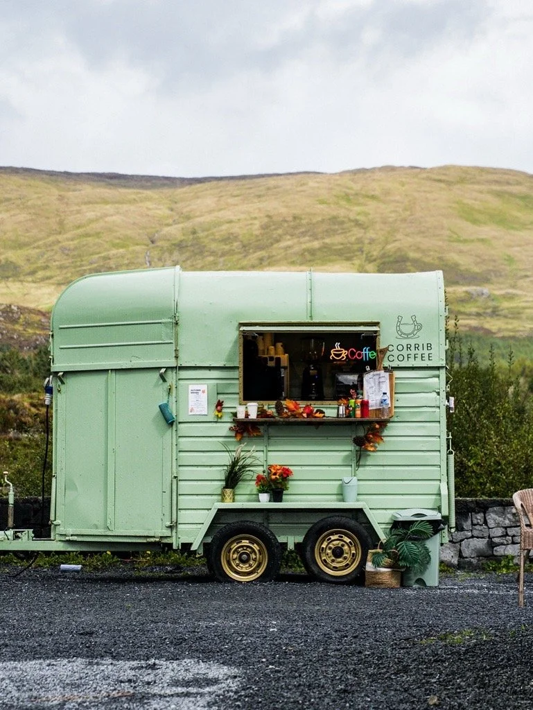 A small mint-green mobile coffee stand with a window serving coffee, decorated with flowers, plants, and signs, set against a grassy hillside and cloudy sky.