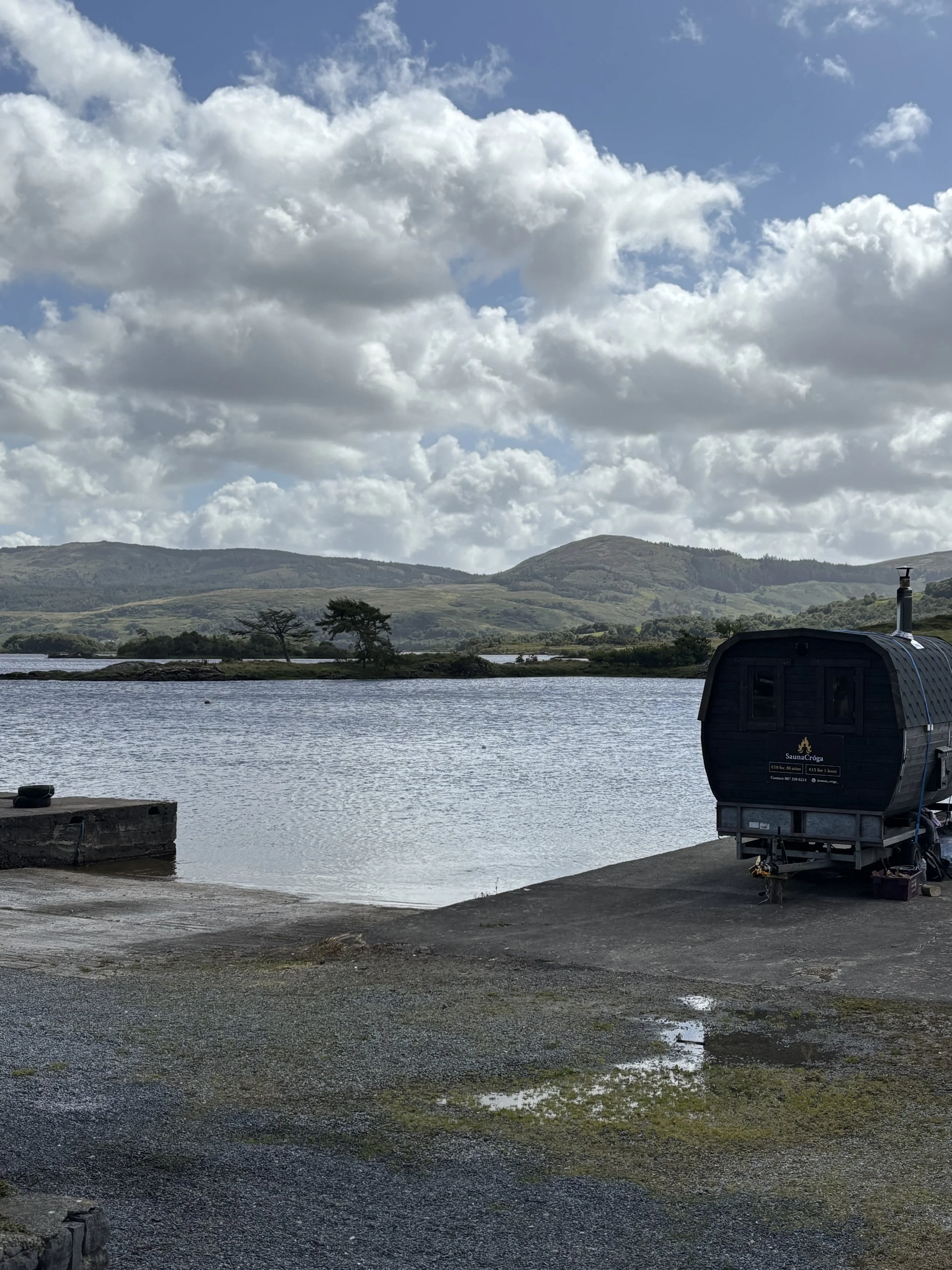 A lake with mountains in the background under a cloudy sky, and a black sauna on wheels on the shore.