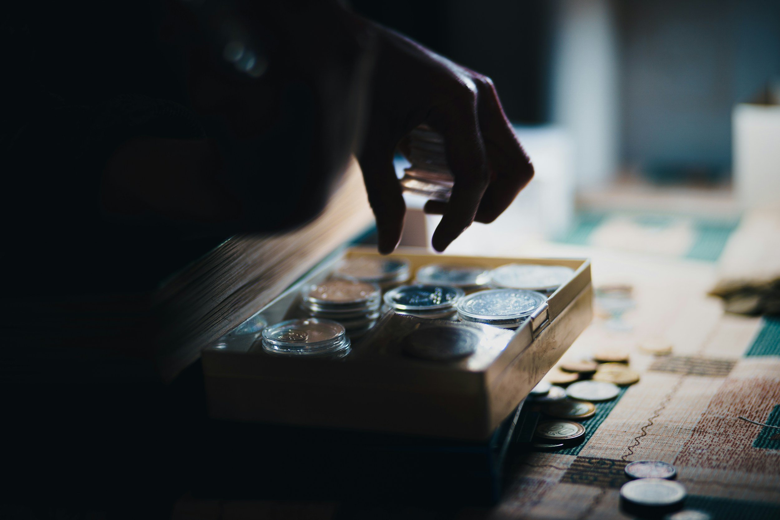 Close-up of a hand reaching into a wooden box filled with coins, with more coins scattered on a table nearby.