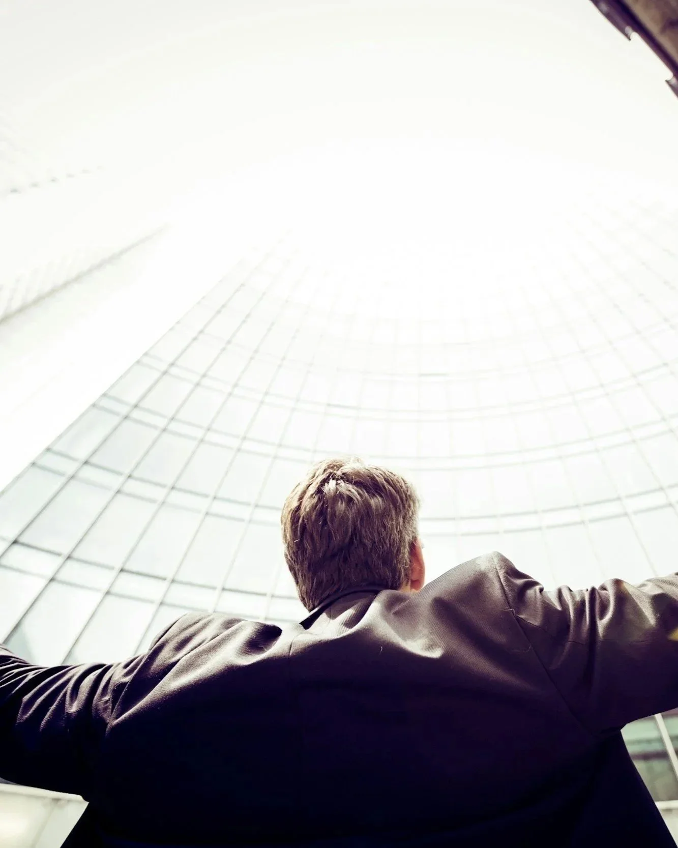 A man with light brown hair wearing a dark jacket, viewed from behind, standing inside a glass-domed building and looking upward toward the bright sky visible through the glass ceiling.