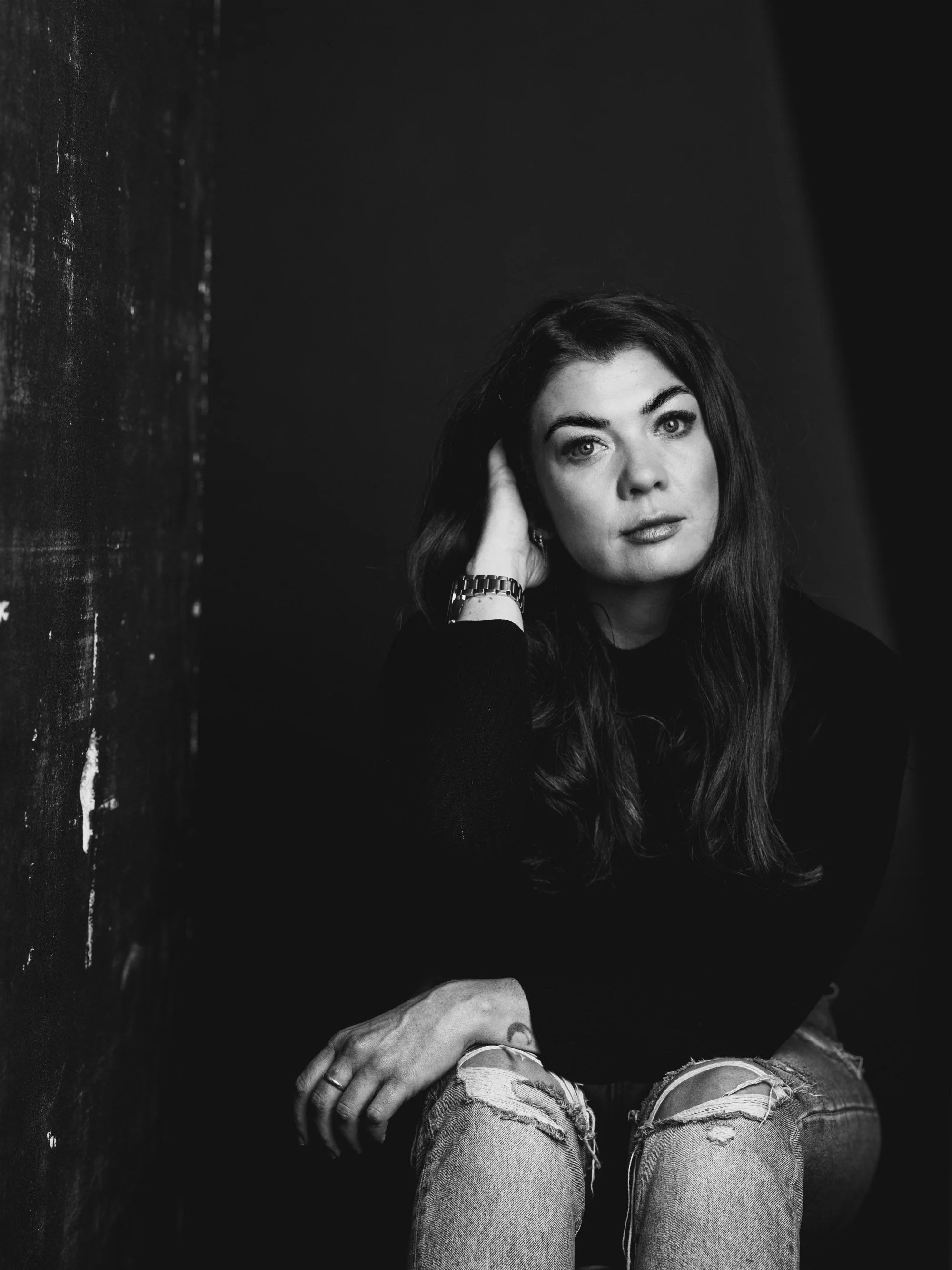 A black and white portrait of a woman with long dark hair, sitting against a dark textured wall. She is wearing a black top, ripped jeans, a watch, and earrings, with her left hand on her head and her right hand resting on her knee.