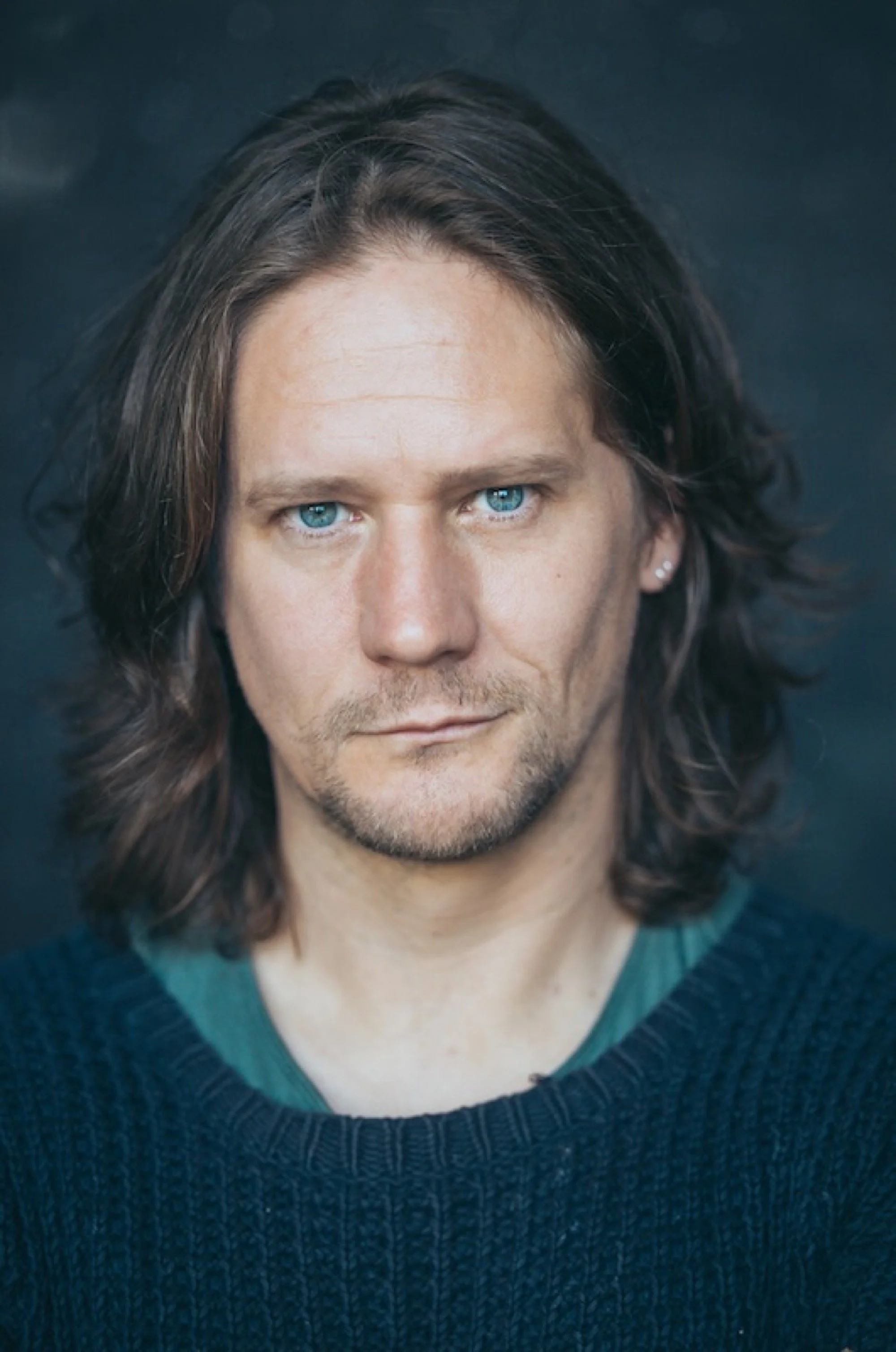 A close-up portrait of a man with long brown hair, blue eyes, and a light beard, wearing a dark sweater.