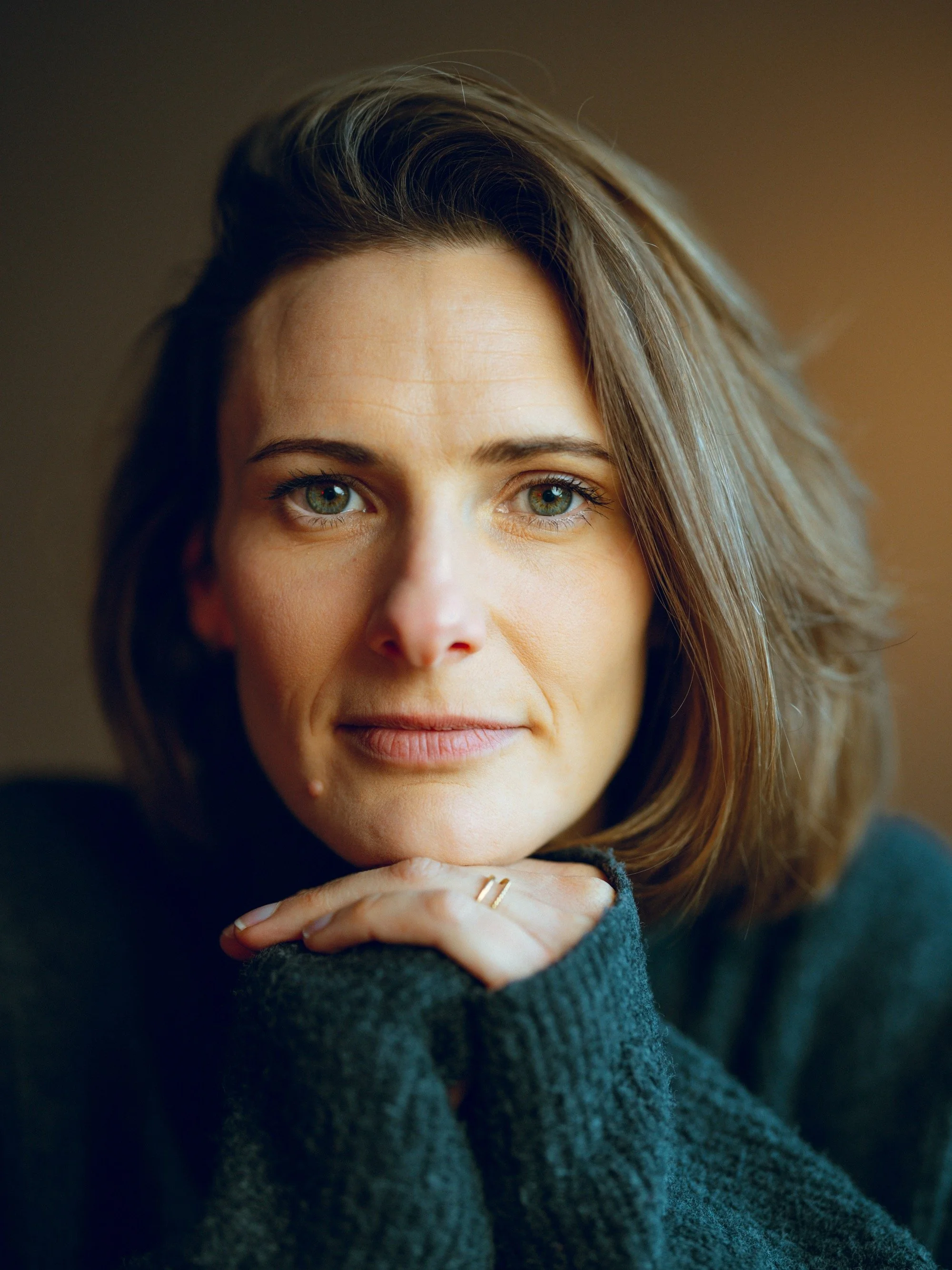 Close-up portrait of a woman with shoulder-length brown hair and blue-green eyes, resting her chin on her folded hands and looking at the camera.