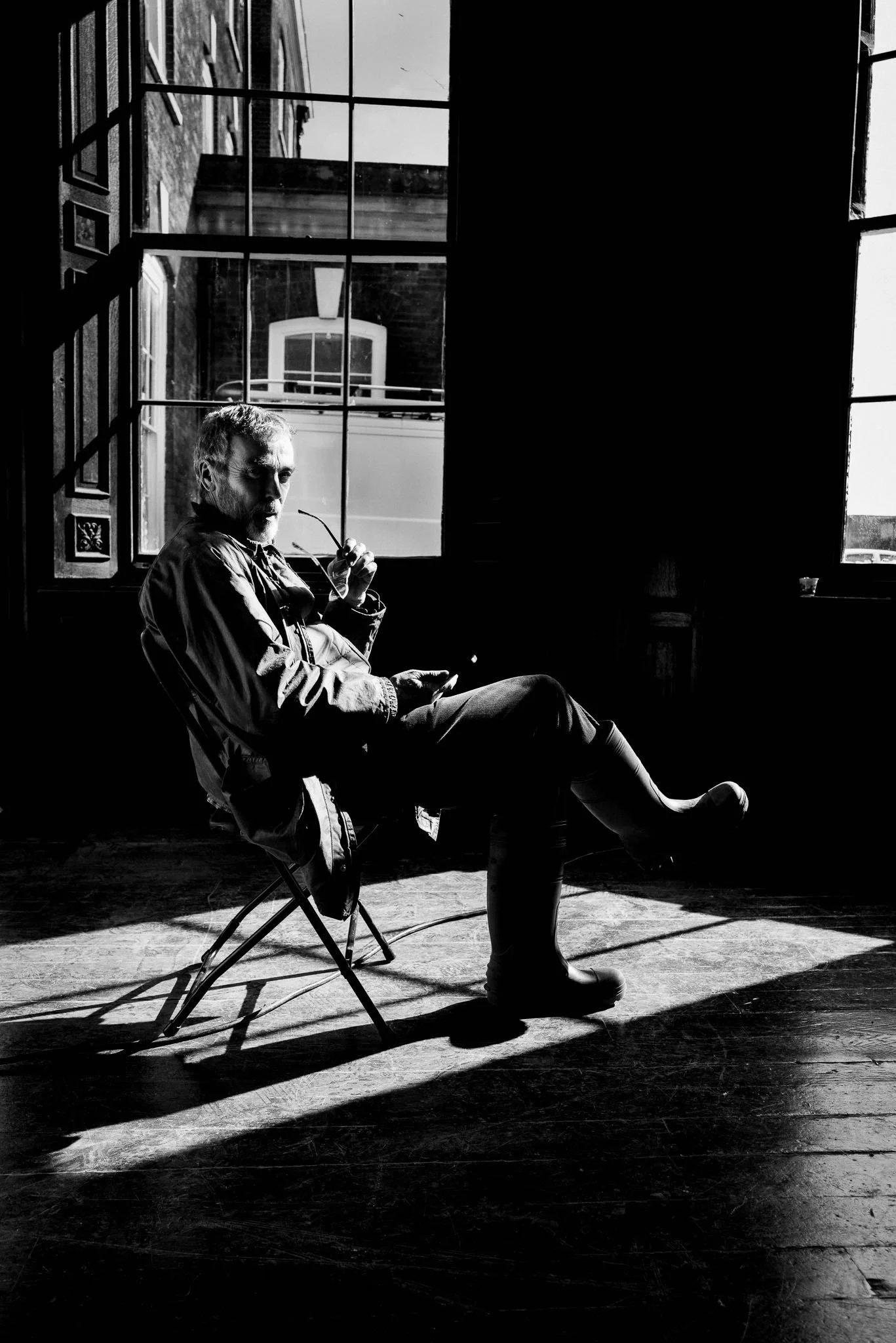 A man sitting on a foldable chair in a room with large windows, holding a pair of glasses and looking towards the camera. Sunlight streams through the windows, casting shadows on the wooden floor.