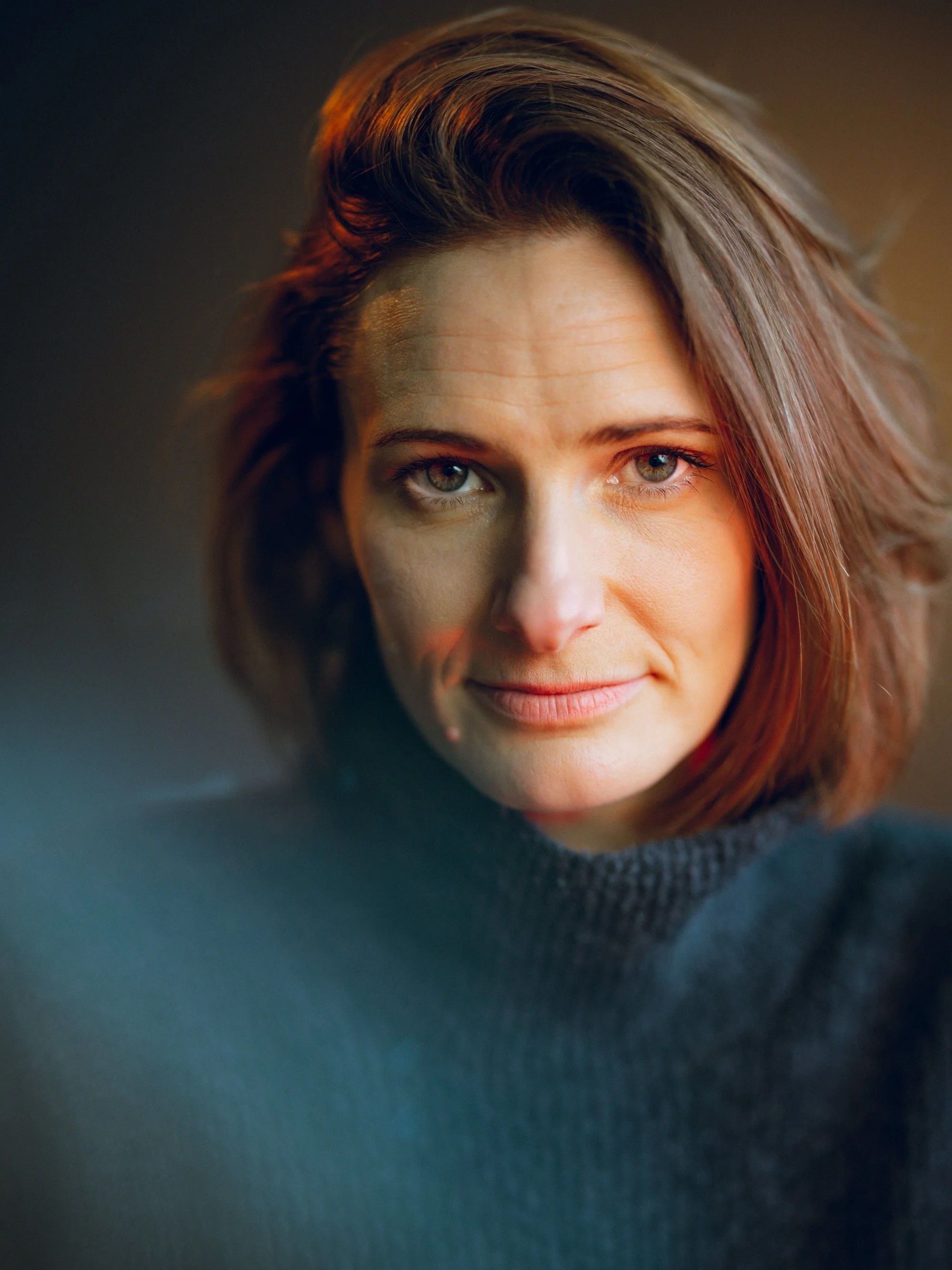 Close-up of a woman with wavy, shoulder-length brown hair and blue eyes, wearing a dark sweater, looking into the camera with a slight smile.