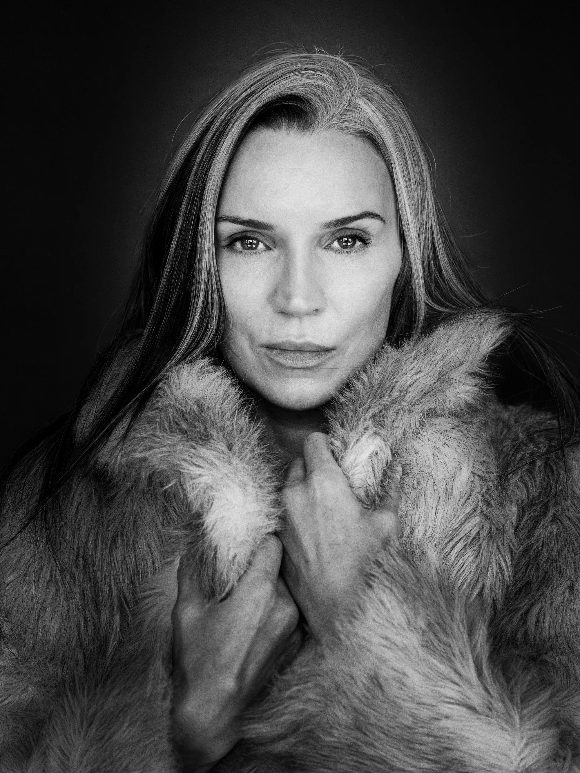Black and white portrait of a woman with light-colored hair, holding a fur coat close to her chest, against a dark background.