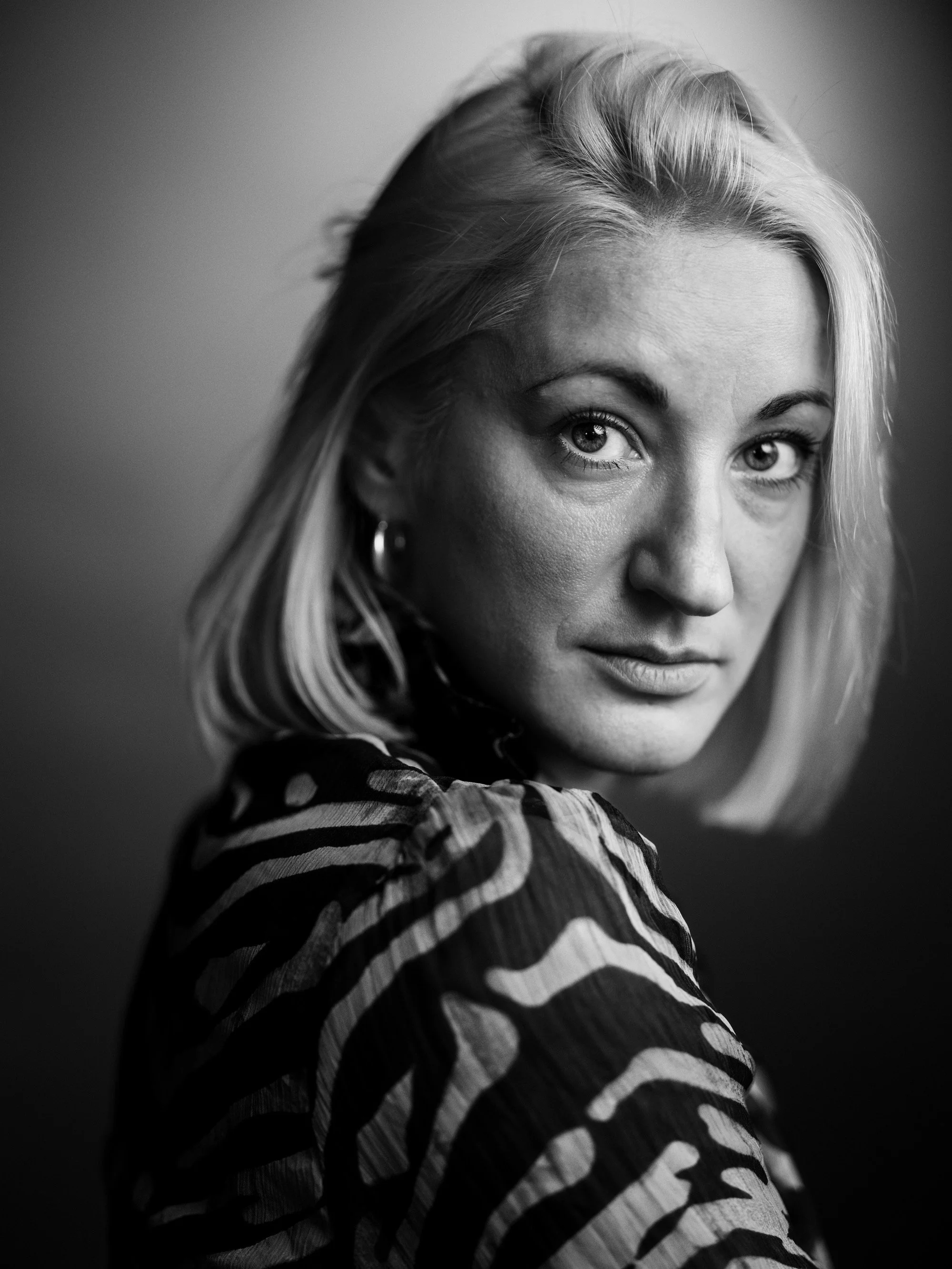 Black and white portrait of a woman with blonde hair, looking at the camera, wearing a patterned top.