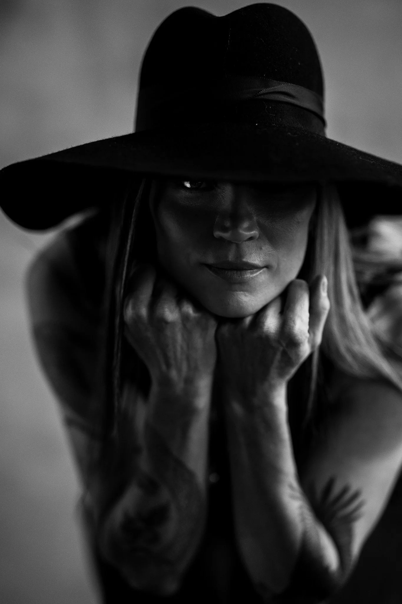 A woman with long hair, wearing a large hat, resting her chin on her hands, looking at the camera in black and white.