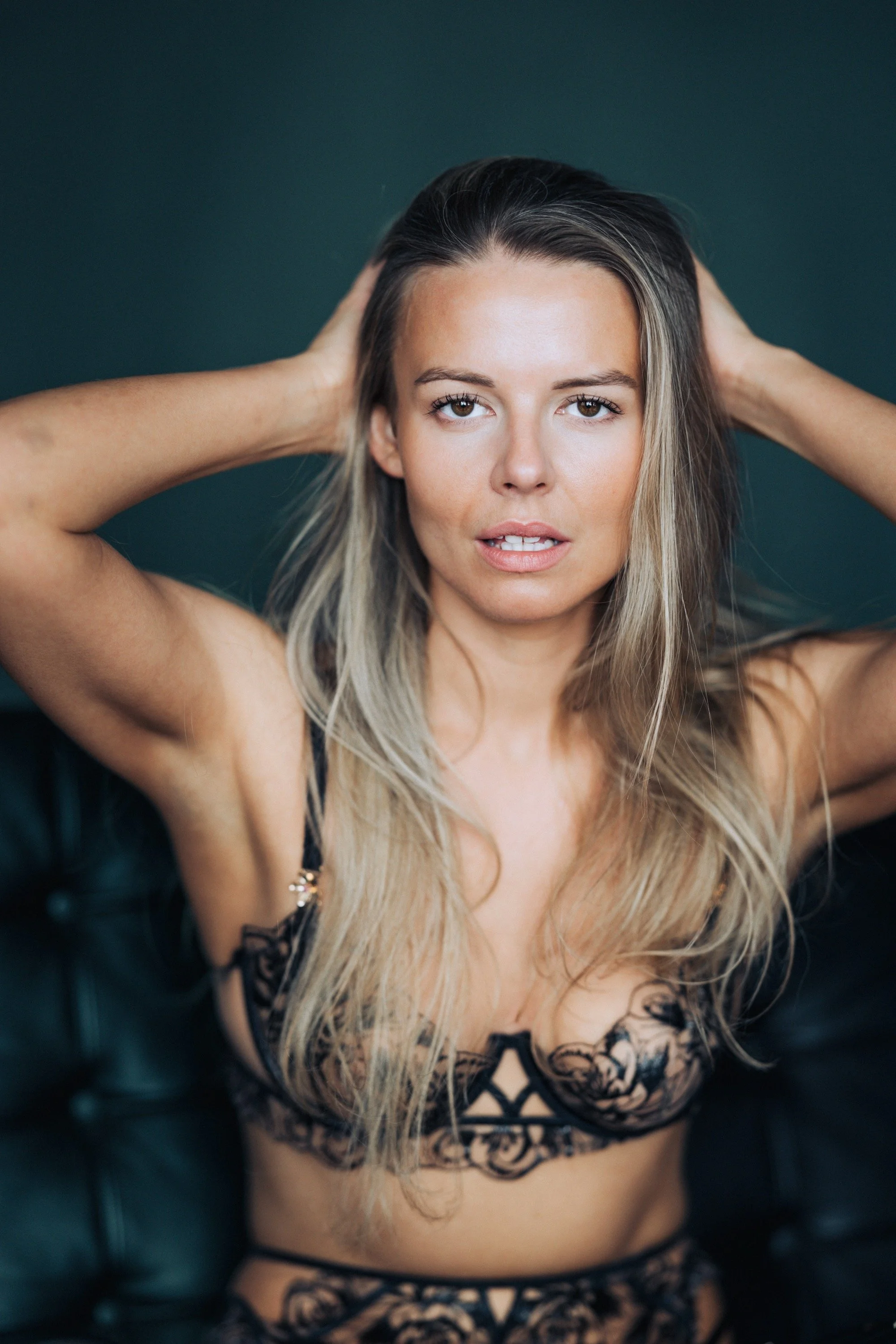 Woman with long blonde hair, wearing black lace lingerie, posing with hands behind her head against dark background.
