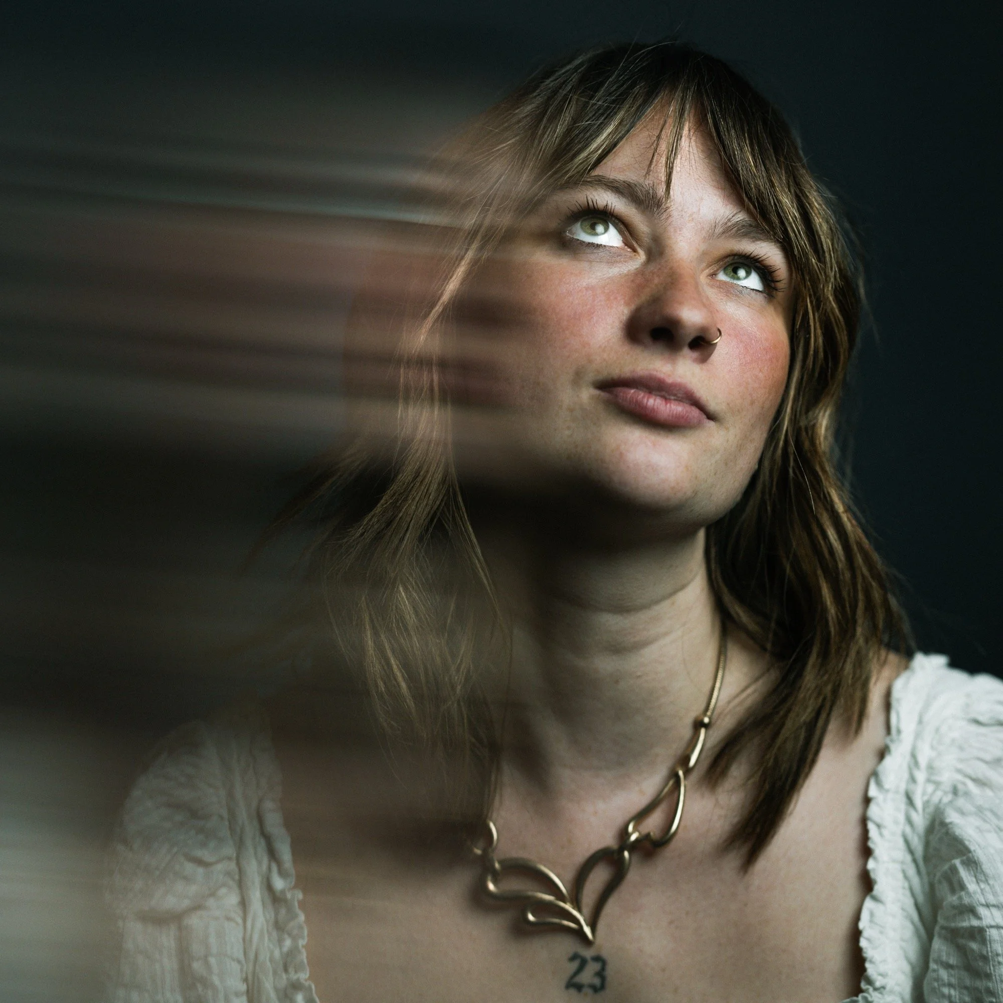 Portrait of a young woman with brown hair, wearing a white top and a silver necklace with a number 23 pendant, looking upwards with light streaks across her face.