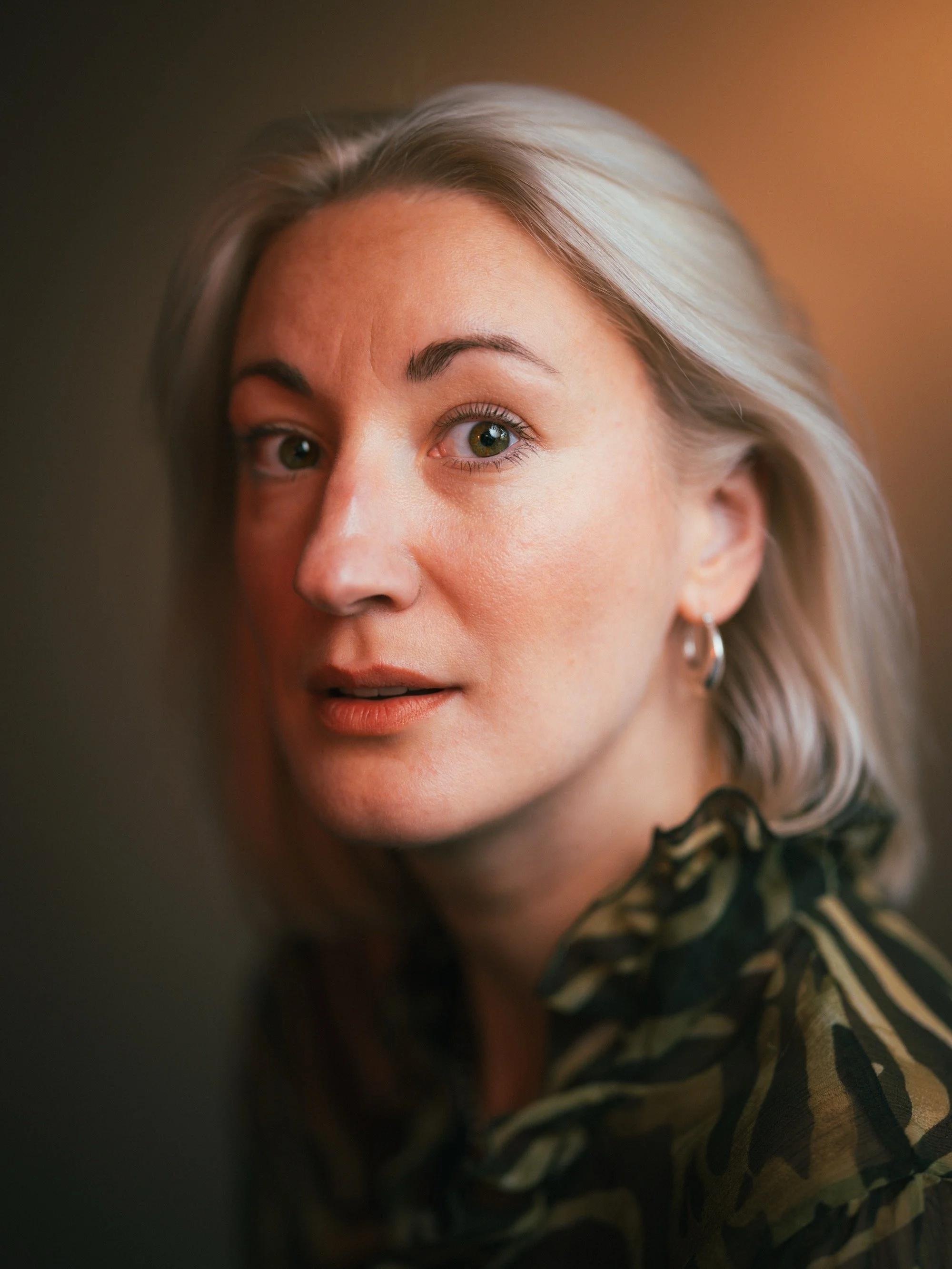 Close-up portrait of a woman with wavy gray hair, wearing a camouflage-patterned shirt and small hoop earrings, looking directly at the camera with a neutral expression.