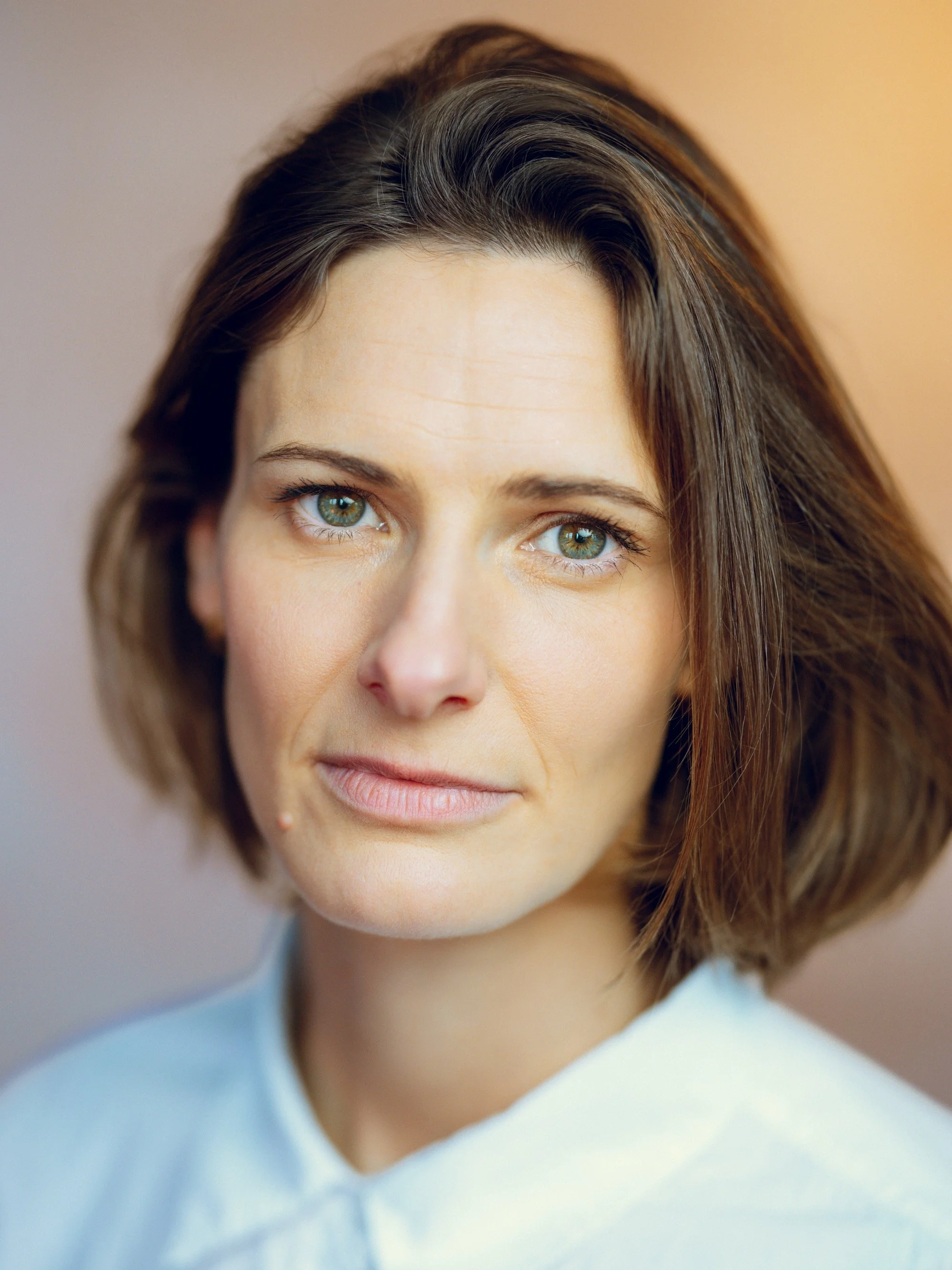 Close-up portrait of a woman with shoulder-length brown hair and green eyes, wearing a light blue shirt.