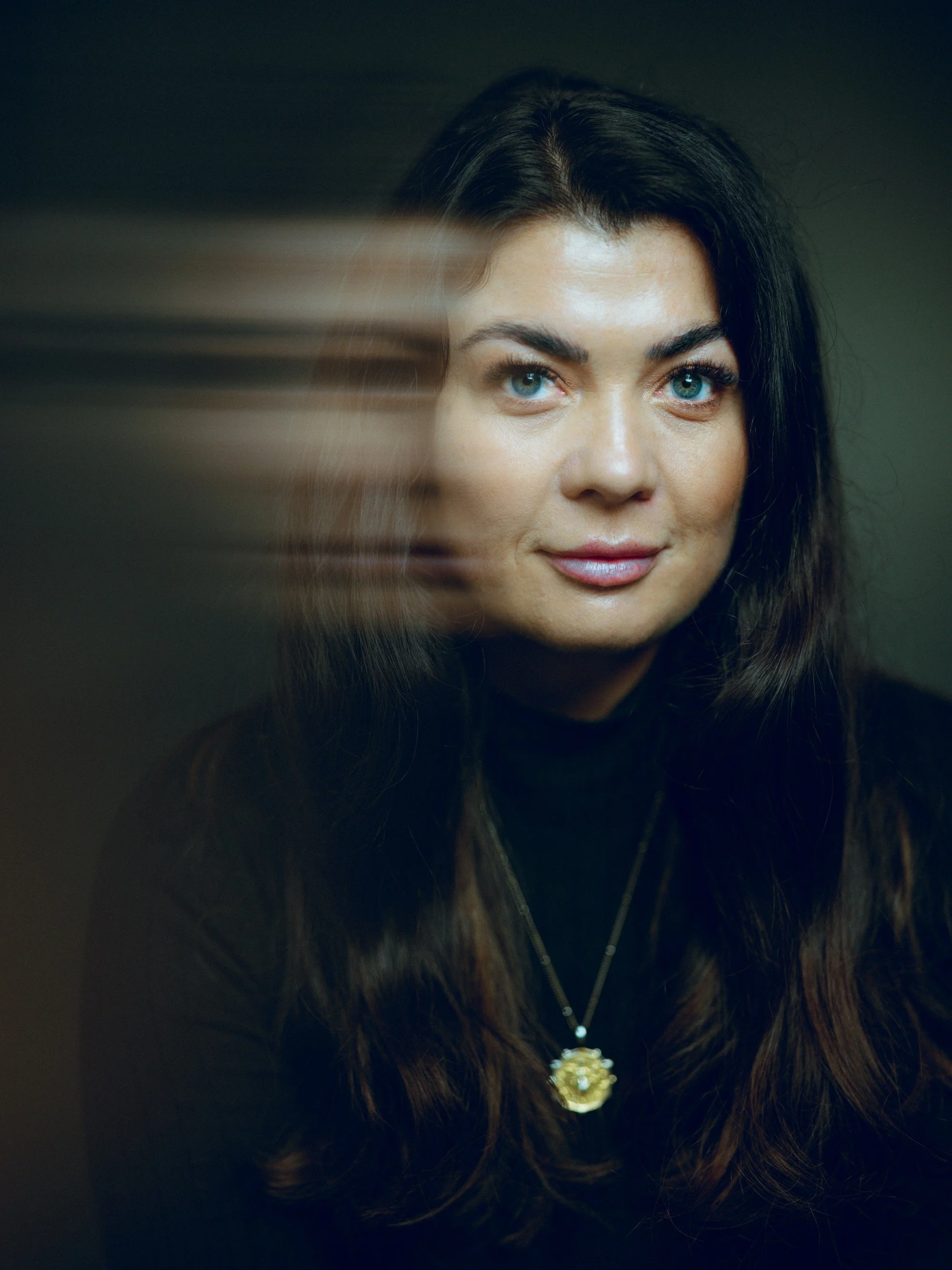 Portrait of a woman with long dark hair, blue eyes, wearing a black turtleneck and a gold pendant necklace, with a blurred motion effect to the left.