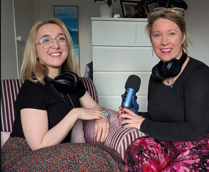 Two women sitting inside a room, both wearing black shirts and headphones around their necks. One woman on the left has blonde hair, glasses, and is sitting on a striped sofa, smiling. The woman on the right has short hair, glasses on her head, and is holding a blue microphone towards the woman on the left. Behind them is a white dresser, a potted plant, and some framed photos or artwork.
