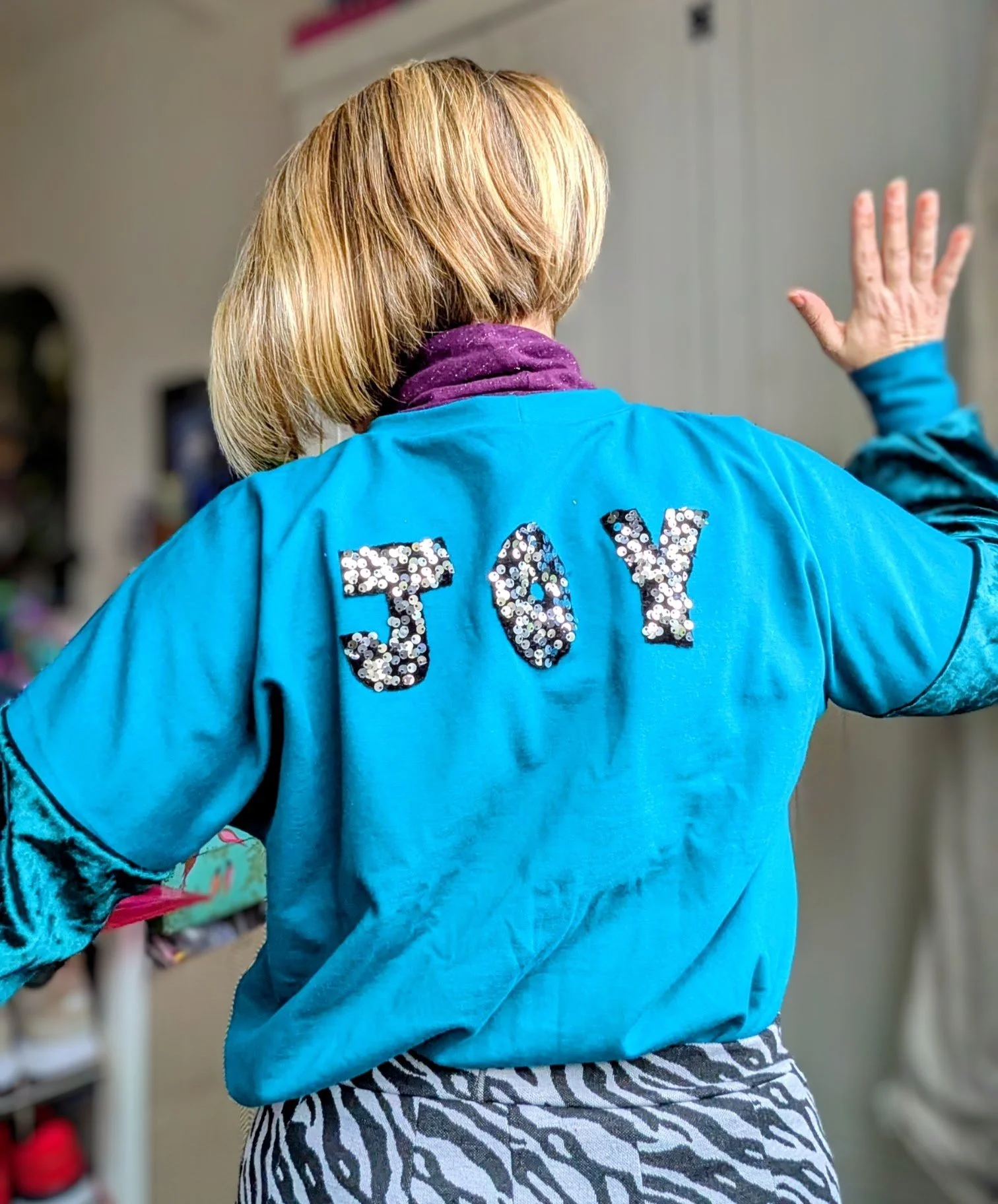 Back view of woman wearing a blue jacket with the word 'JOY' in sequined letters, waving her right hand inside a room.