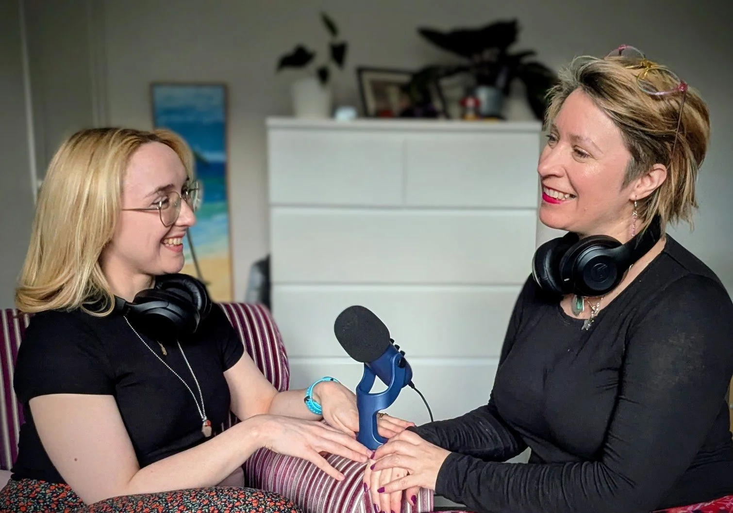 Two women smiling and holding hands during an interview or podcast recording, with a microphone between them and headphones around their necks.