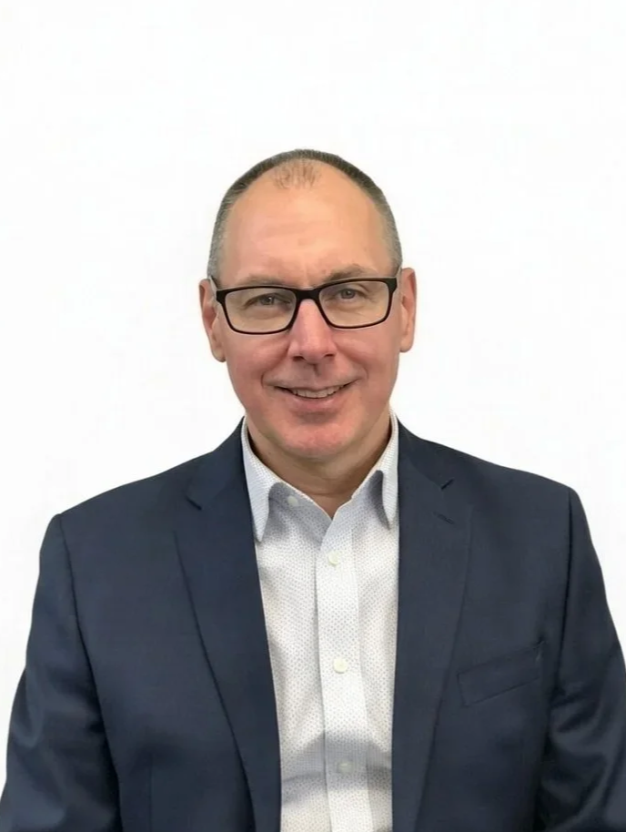 An executive with glasses, short hair, wearing a dark suit jacket and white shirt, smiling against a plain white background.