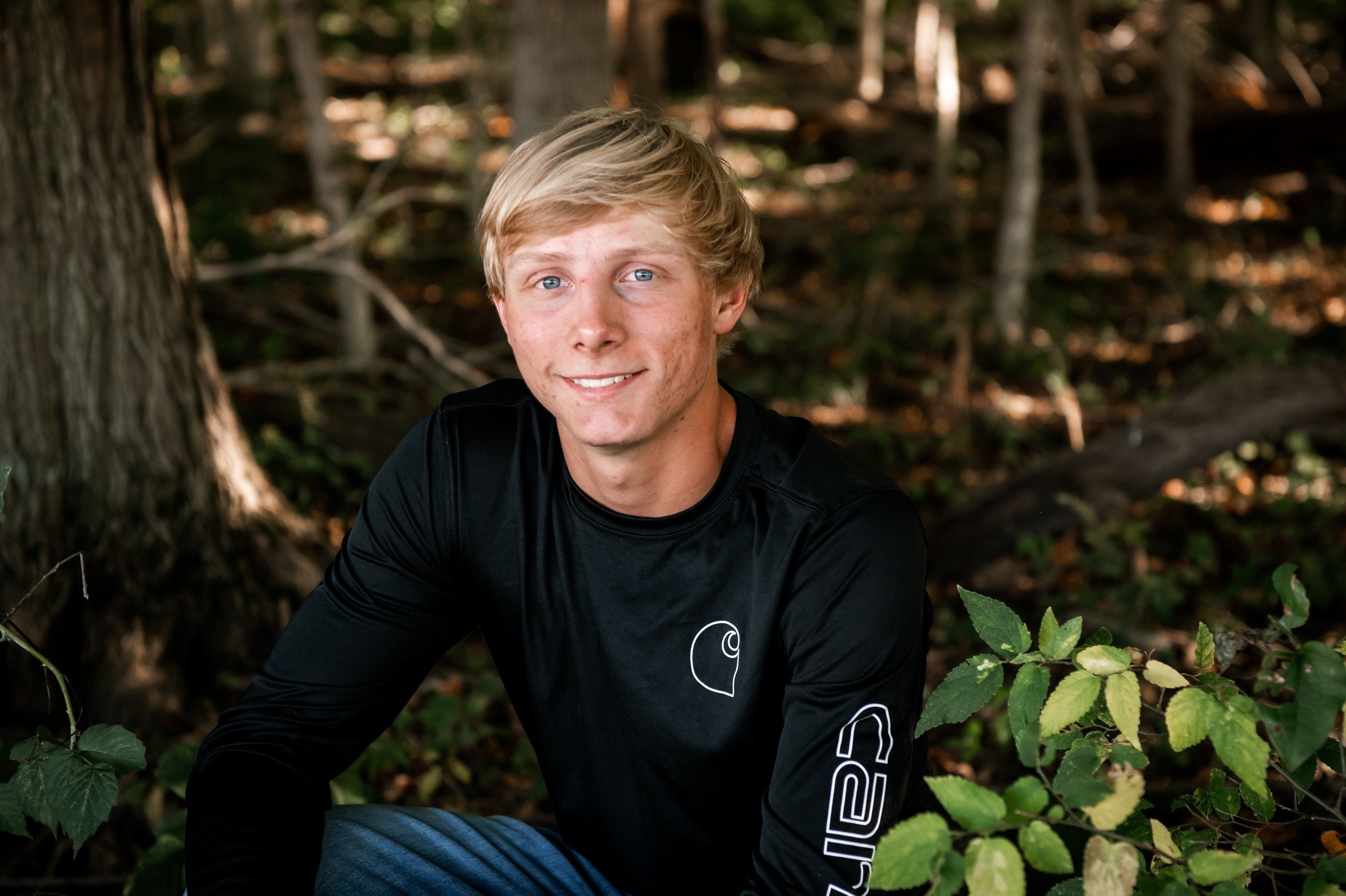 A young man with blond hair and blue eyes, smiling, wearing a black long-sleeve shirt, sitting outdoors in a wooded area with trees and green foliage.