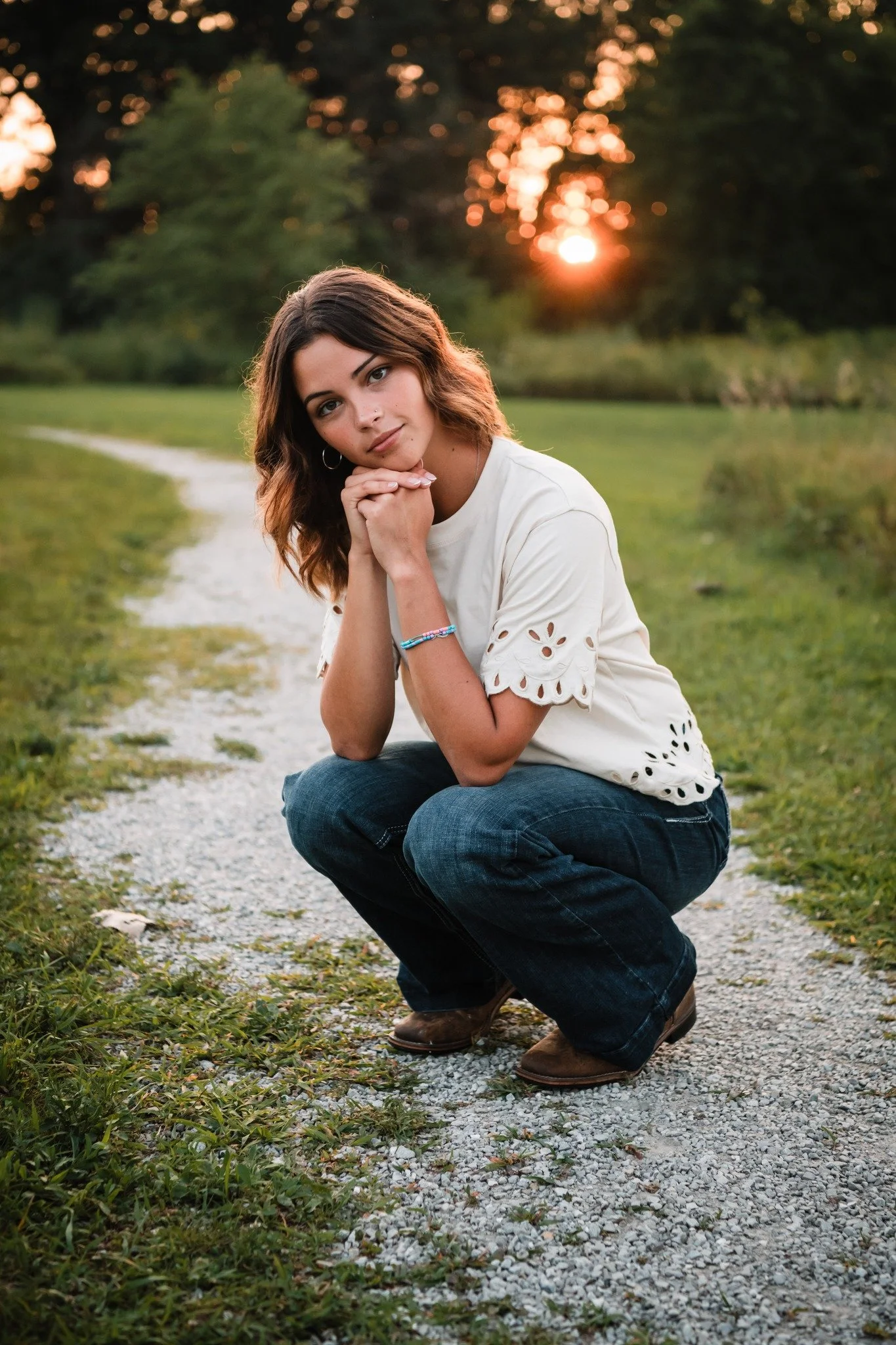A young woman with wavy brown hair wearing a white top and dark blue jeans is squatting on a gravel path in a grassy park during sunset, with her hands clasped under her chin and looking at the camera.