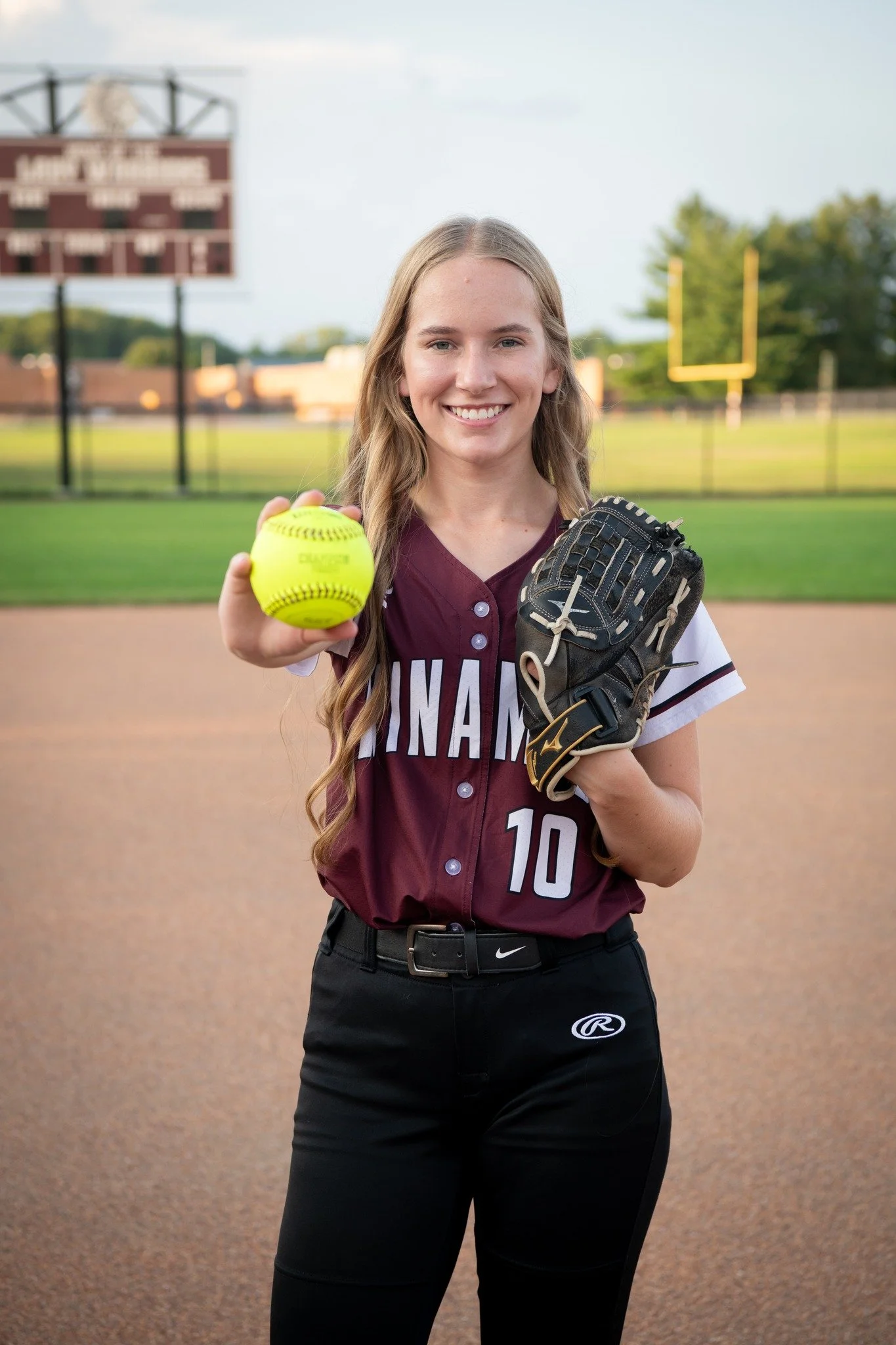Young woman in a baseball uniform holding a yellow softball and a baseball glove, standing on a baseball field.