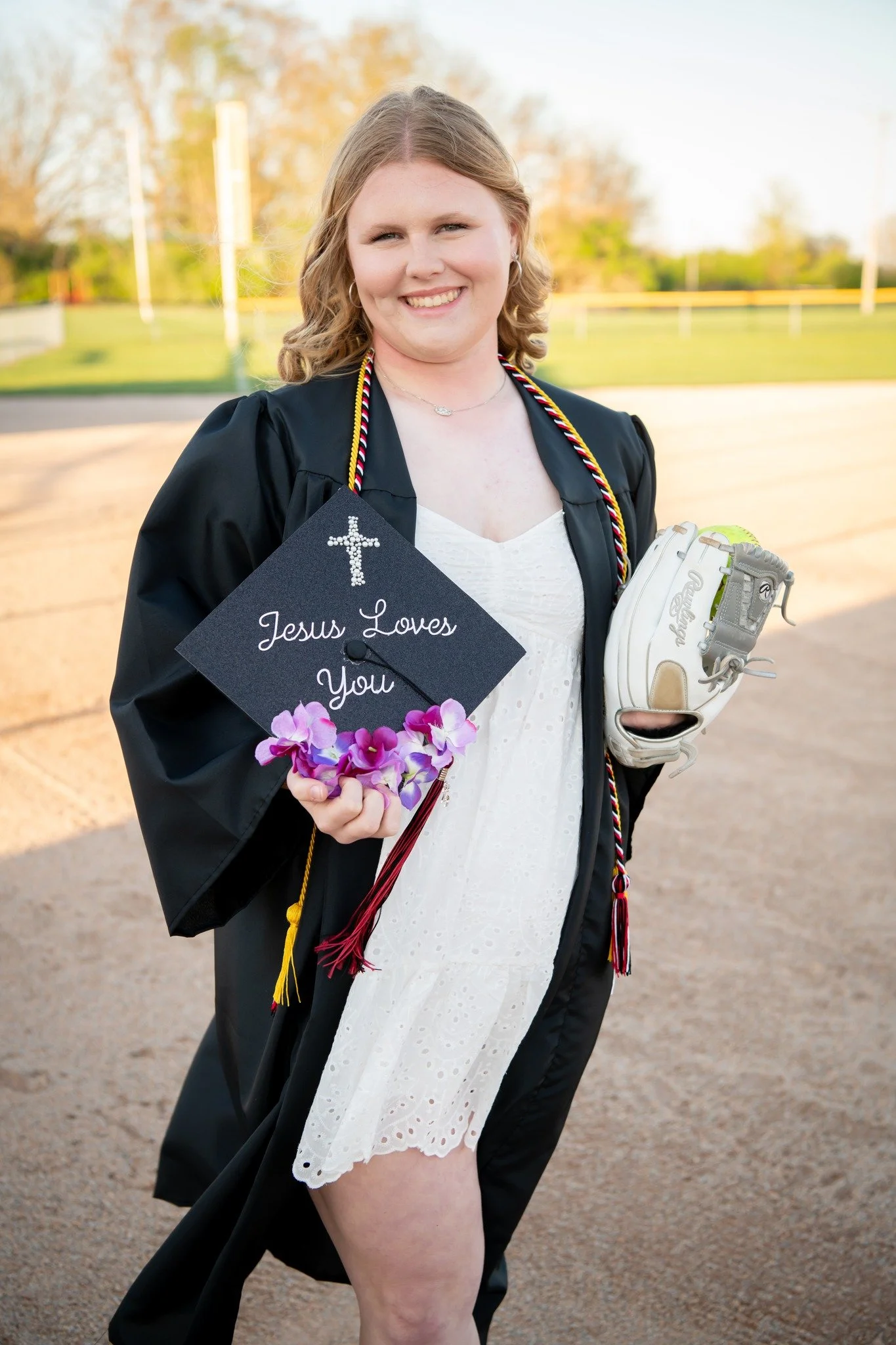 A young woman in a graduation gown holding a sign that says 'Jesus Loves You' on a baseball field with trees in the background.