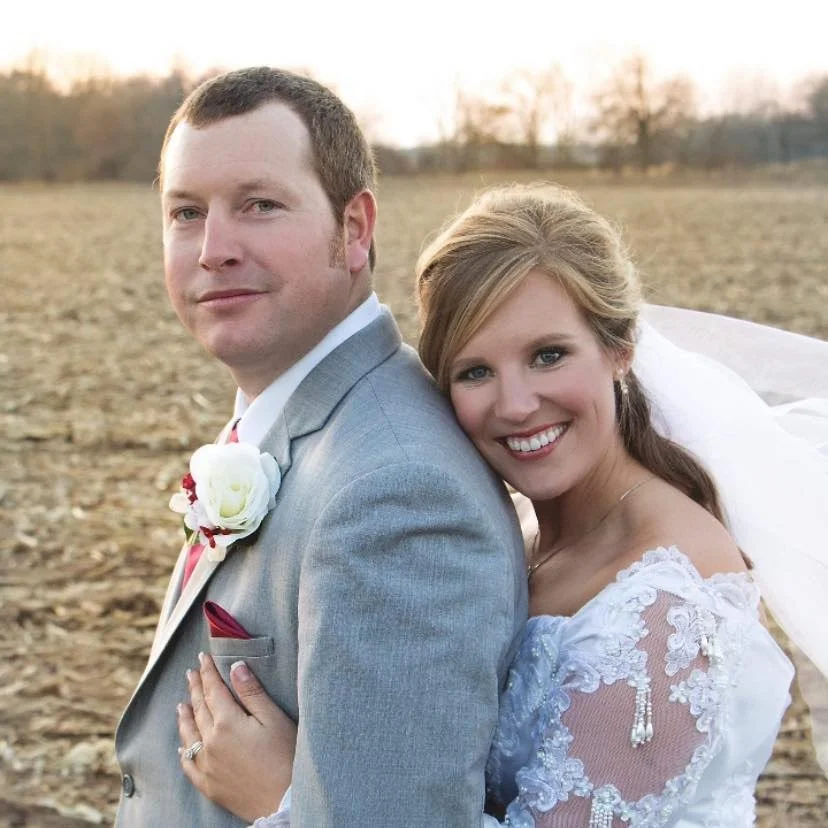 A wedding photo of a smiling bride and groom outdoors in a field during sunset, with the bride leaning on the groom's shoulder.