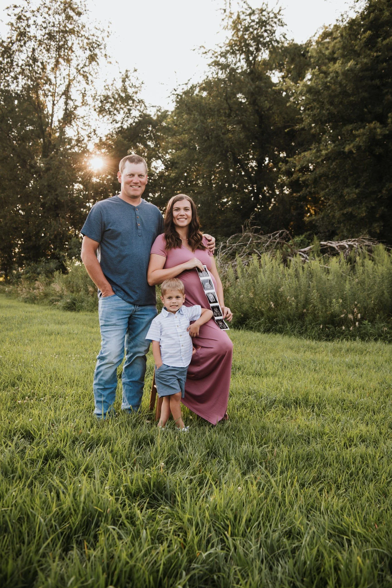 A family of three posing outdoors in a grassy field during sunset, with a pregnant woman holding ultrasound pictures and a young boy standing in front of her.