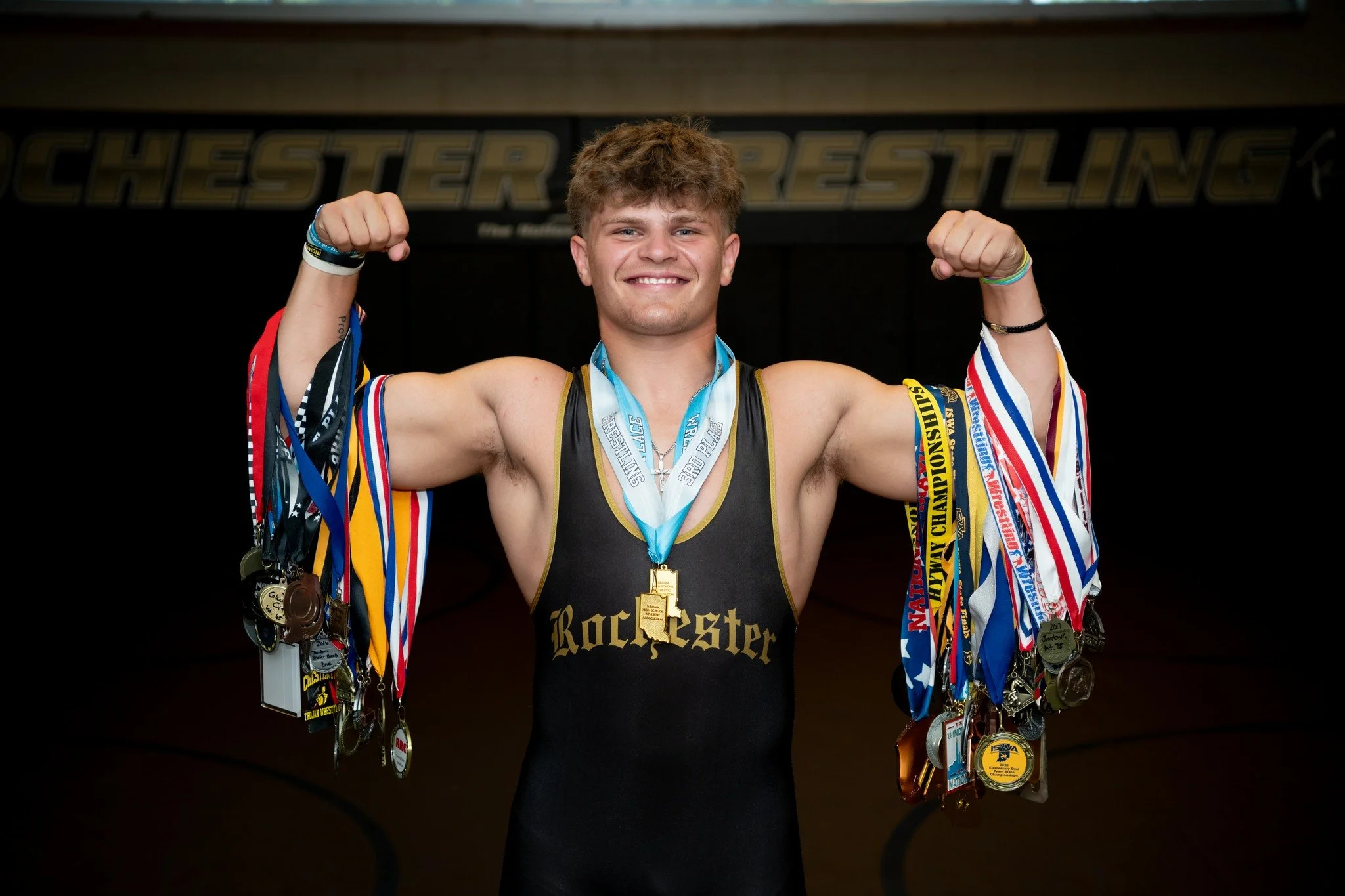 Young male wrestler with medals around his neck, flexing his muscles, wearing a black singlet with 'Rochester' written on it, smiling, with a dark wrestling mat and a banner in the background.