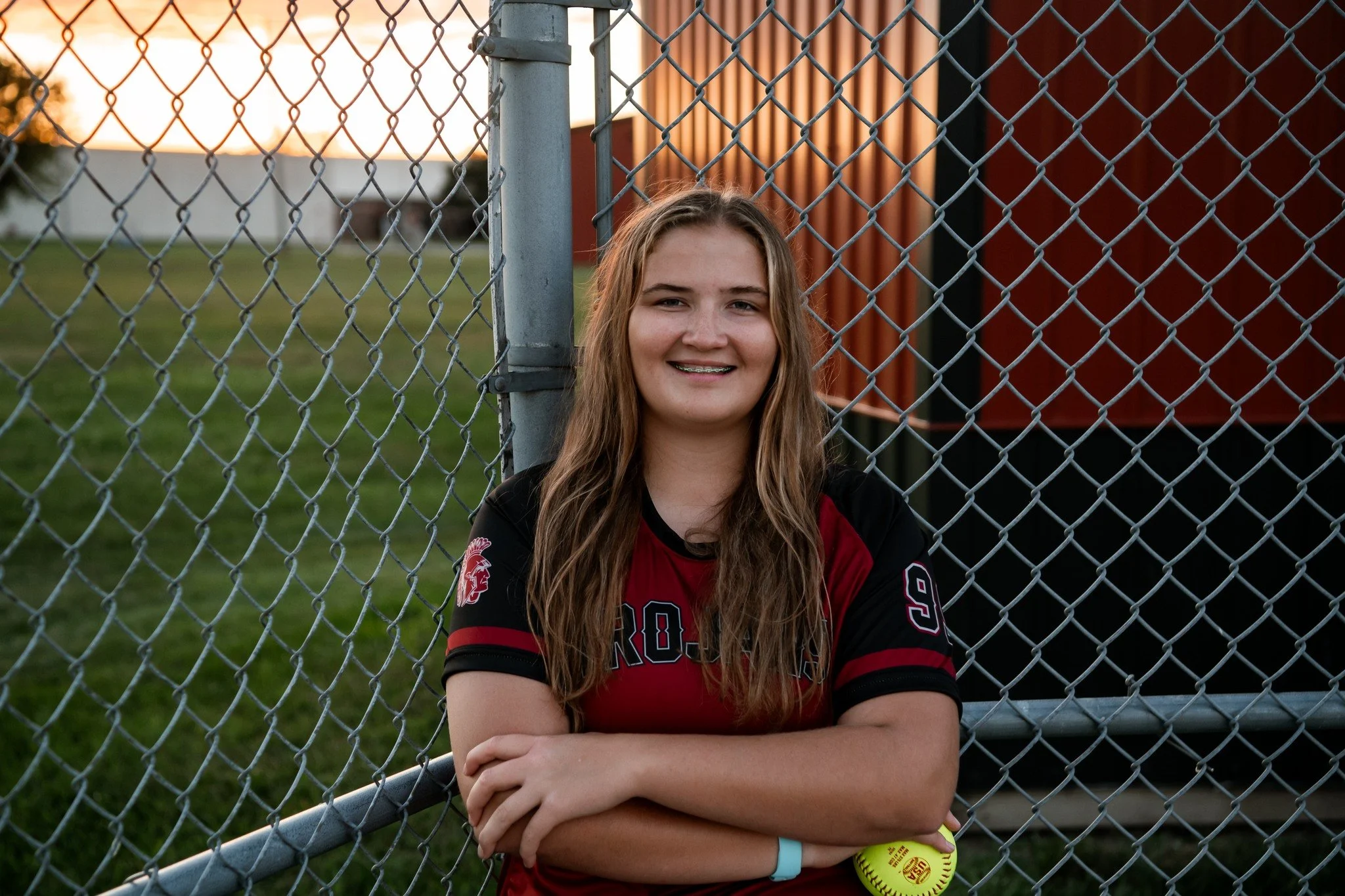 A young female softball player with long hair and braces, wearing a red and black jersey, standing with arms crossed holding a yellow softball and smiling in front of a chain-link fence during sunset.