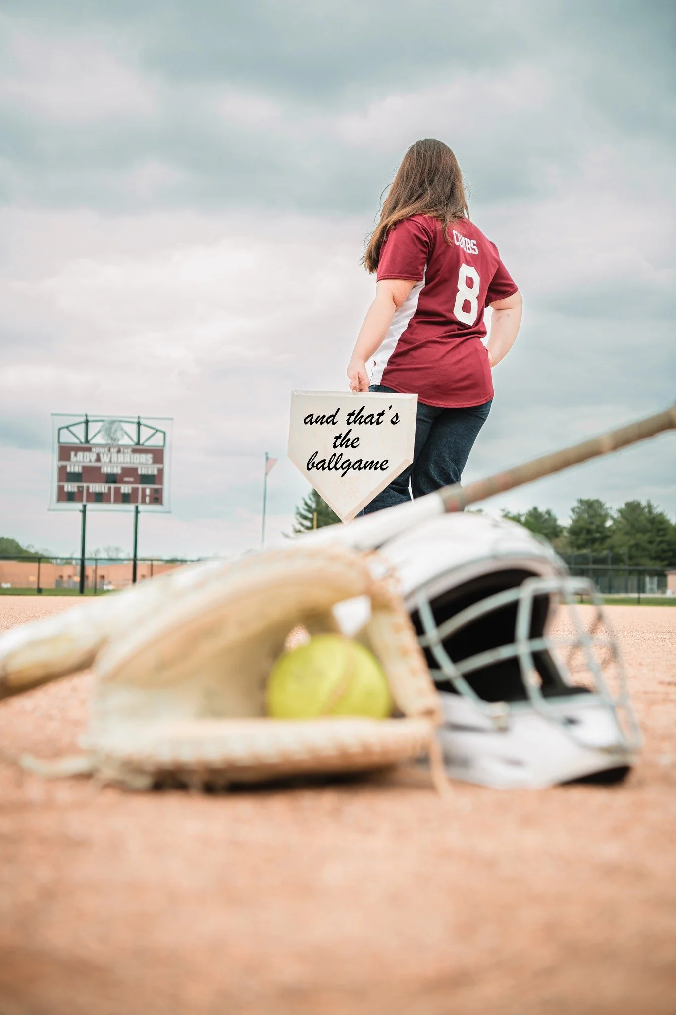 A girl in a sports jersey holding a sign that says 'and that's the ballgame' standing on a baseball field. In the foreground, there is a baseball, a baseball glove, and a catcher's helmet on the ground. The background shows a scoreboard and cloudy sk