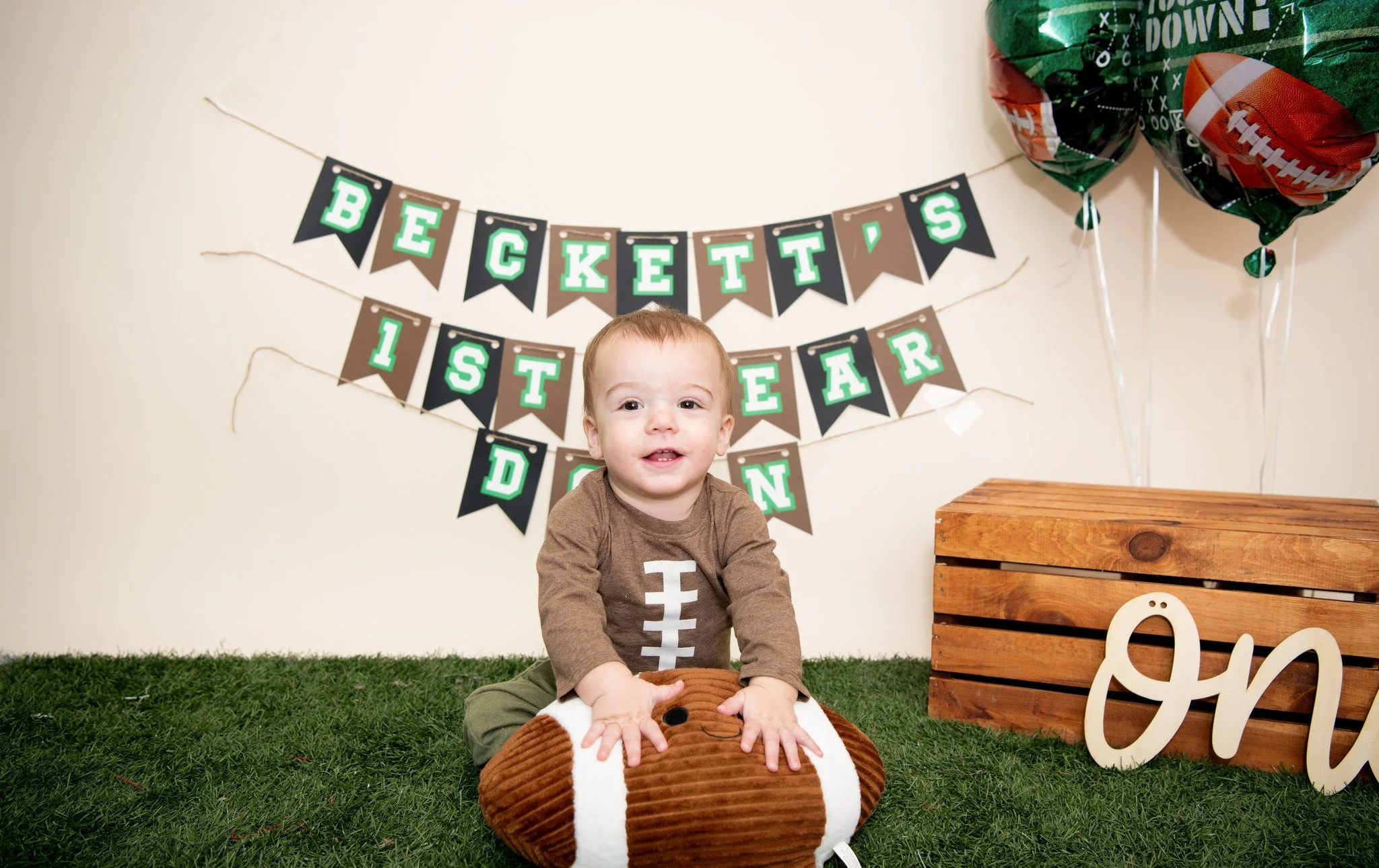 Child celebrating first year birthday in football-themed decorations with 'Beckett's 1st Year' banner, two football balloons, and a wooden crate with a decorative word on it.