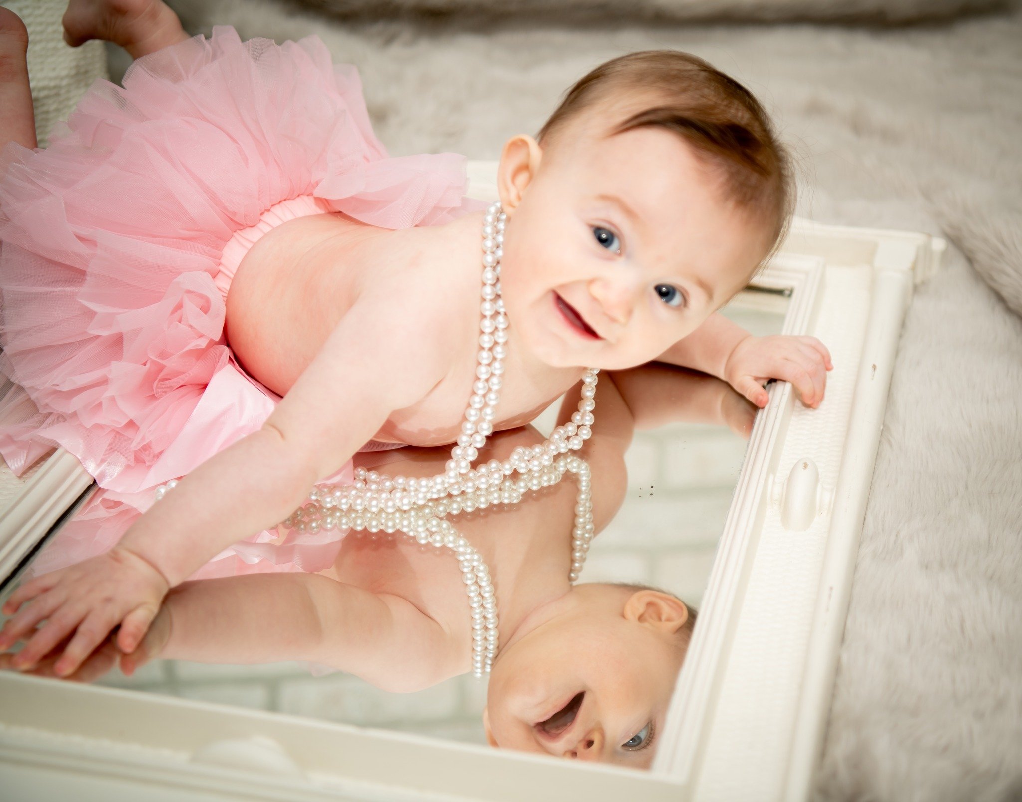 A baby girl in a pink tutu dress and pearl necklace is lying on a mirror, looking up and smiling at the camera.