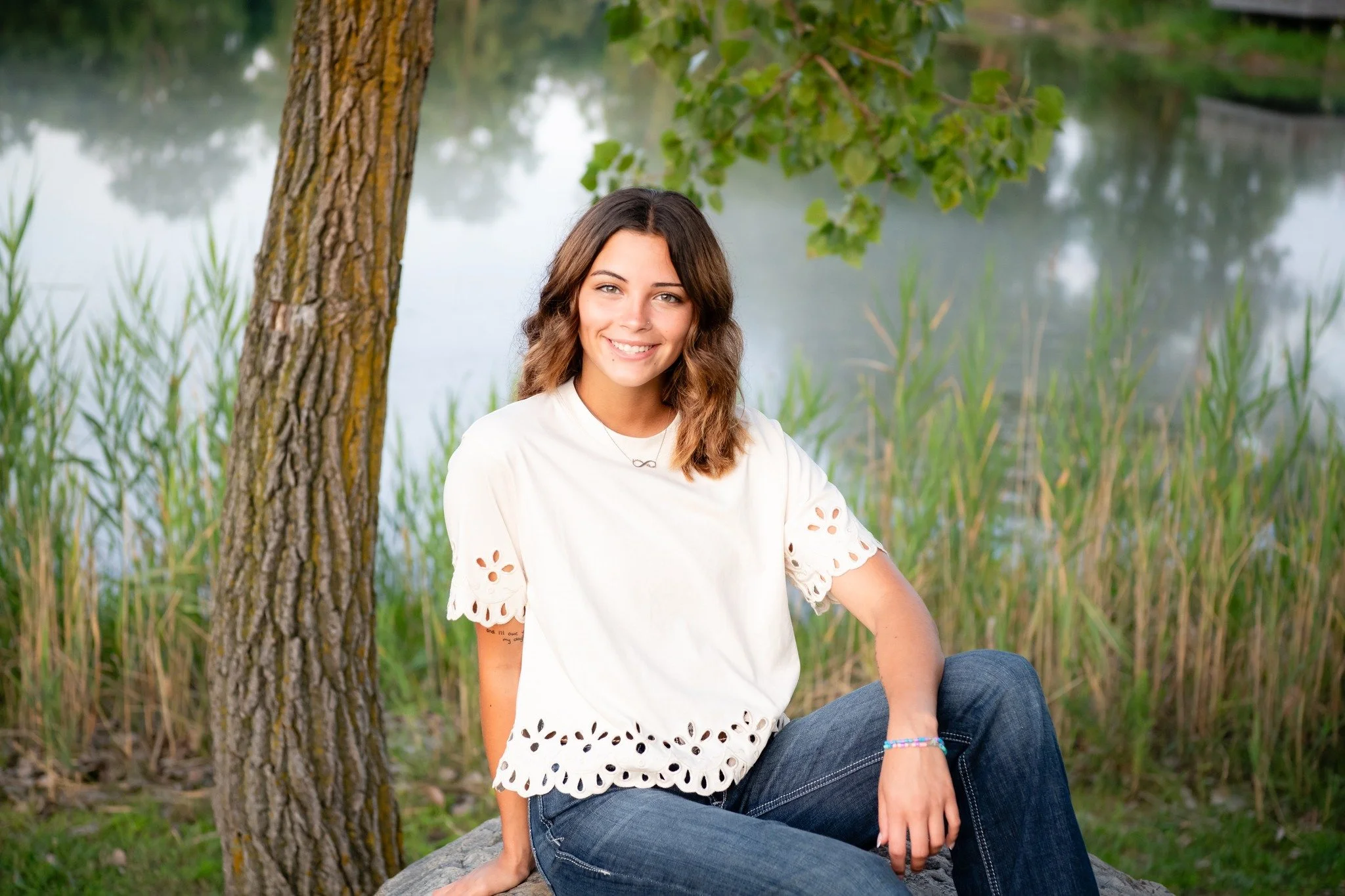 Young woman with wavy brown hair smiling, sitting outdoors by a tree near a body of water, wearing a white eyelet top and blue jeans.