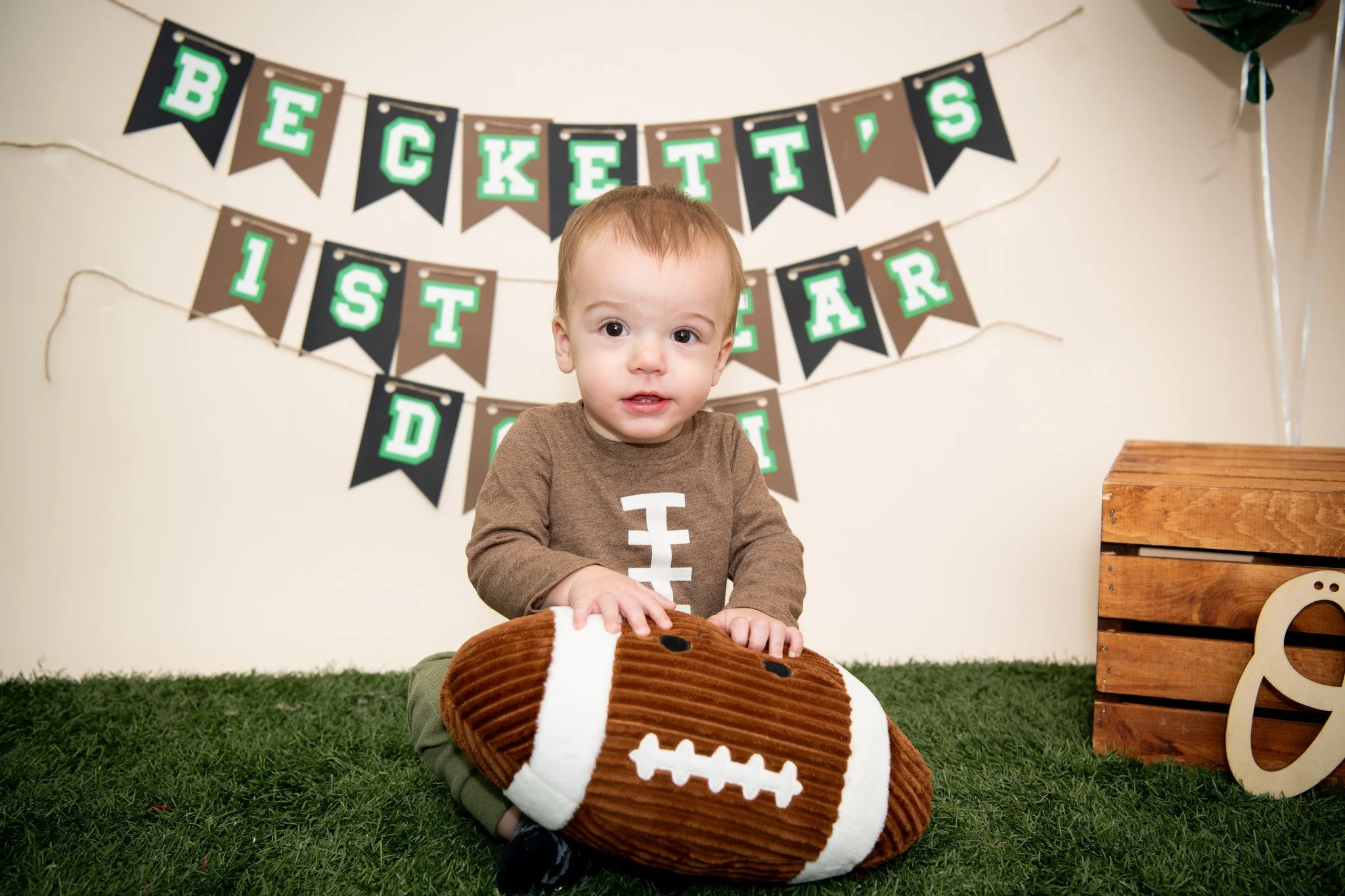 A young boy dressed in a brown football jersey, sitting on grass with a plush football toy, in front of a sign that reads 'Beckett's 1st Year Party'.