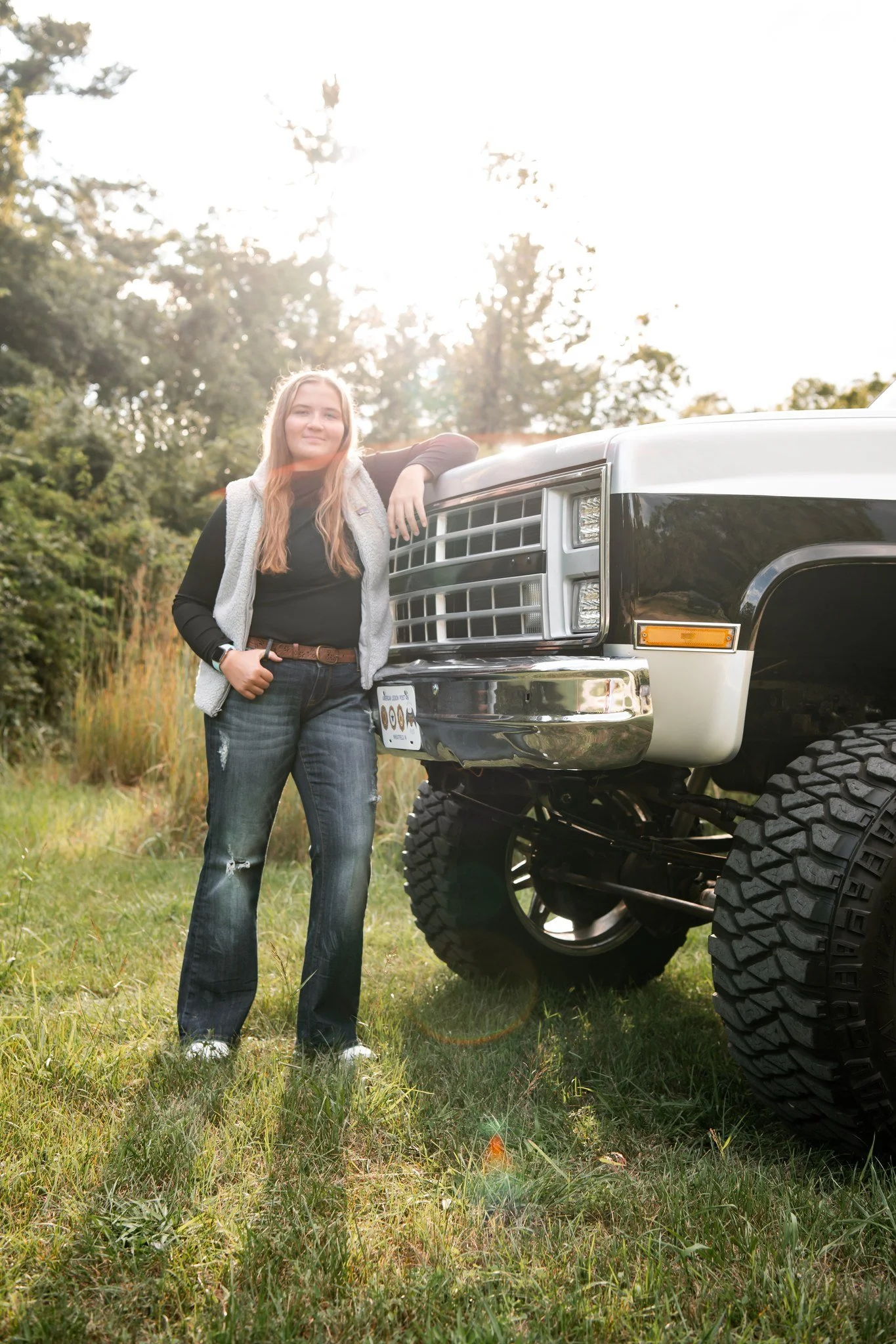 A young woman leaning against a large, lifted black and white truck in a grassy outdoor setting with trees in the background, sunlight filtering through the trees.