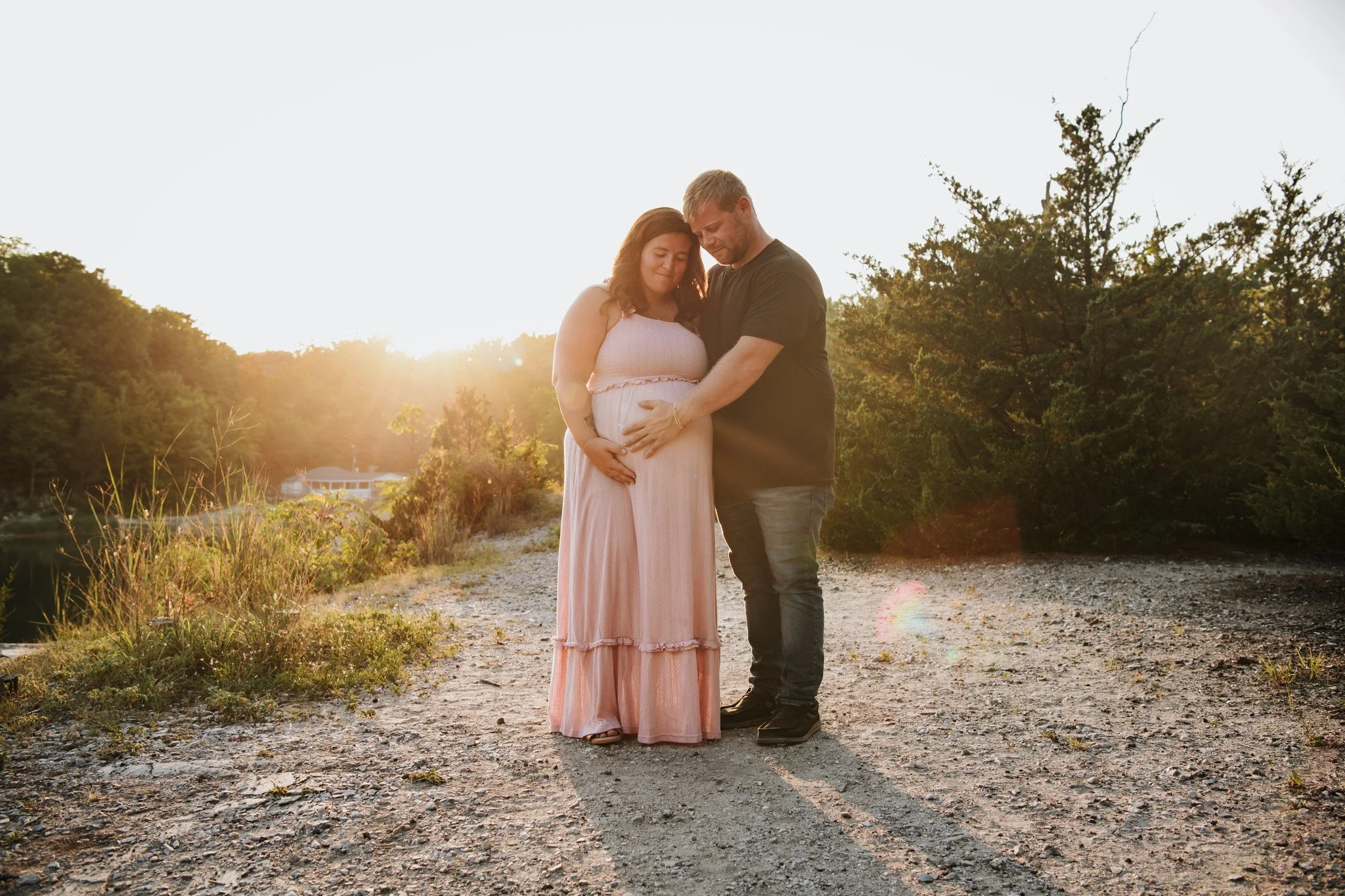 A pregnant woman and a man stand together outdoors during sunset, gently touching her belly and looking at it lovingly, with trees and a clear sky in the background.