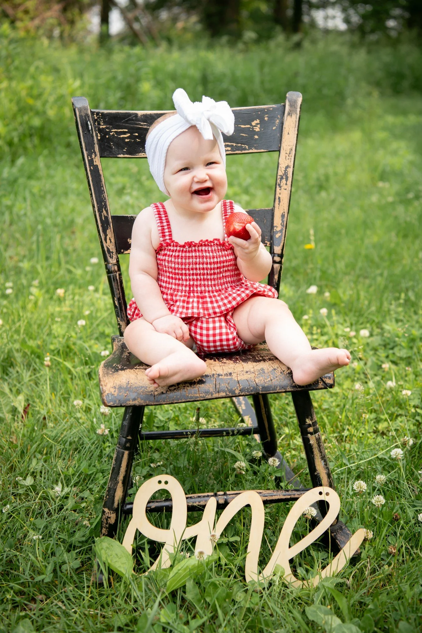 A smiling baby girl with a white headband, wearing a red and white checkered outfit, sitting on an old wooden chair outdoors on grass. She is holding a red apple in her hand, and there is a wooden decorative word 'love' on the grass in front of her.