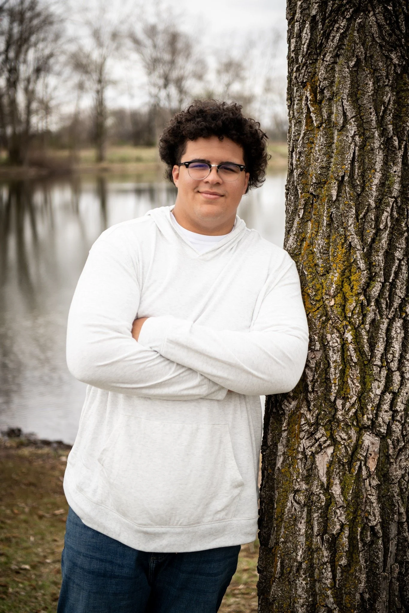 Young man with curly hair and glasses standing outdoors by a large tree near a body of water, arms crossed, wearing a light-colored hoodie.