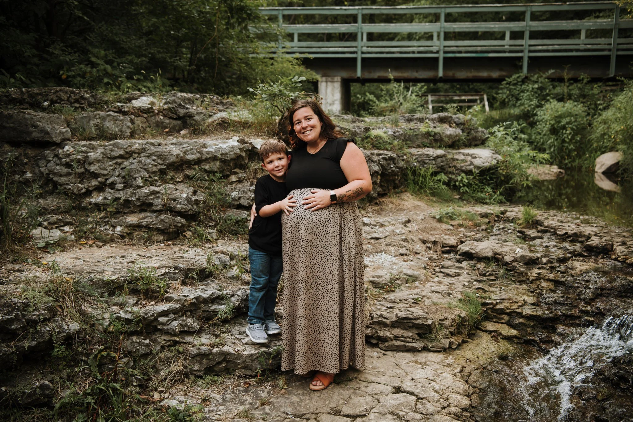 A woman with a pregnant belly and a young boy standing on rocks beside a creek in a green outdoor setting, smiling at the camera.