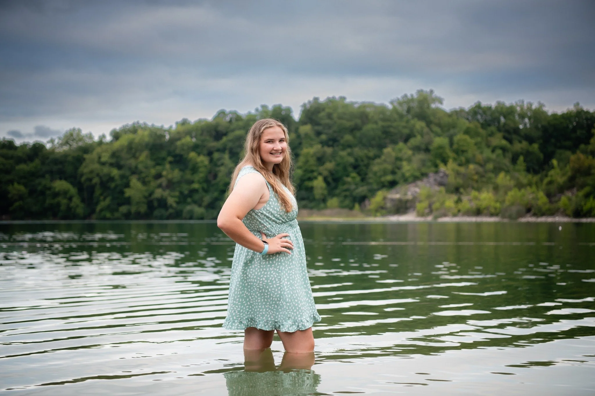 A young woman in a light blue dress standing in a body of water with a forested hill and cloudy sky in the background.