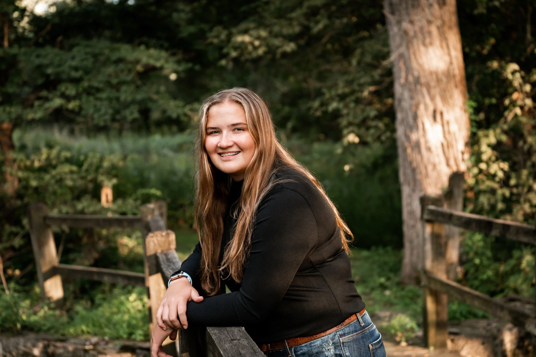 A young woman with long blonde hair, wearing a black long-sleeve shirt and jeans, leaning on a wooden fence in a lush, green outdoor forest setting during daytime.
