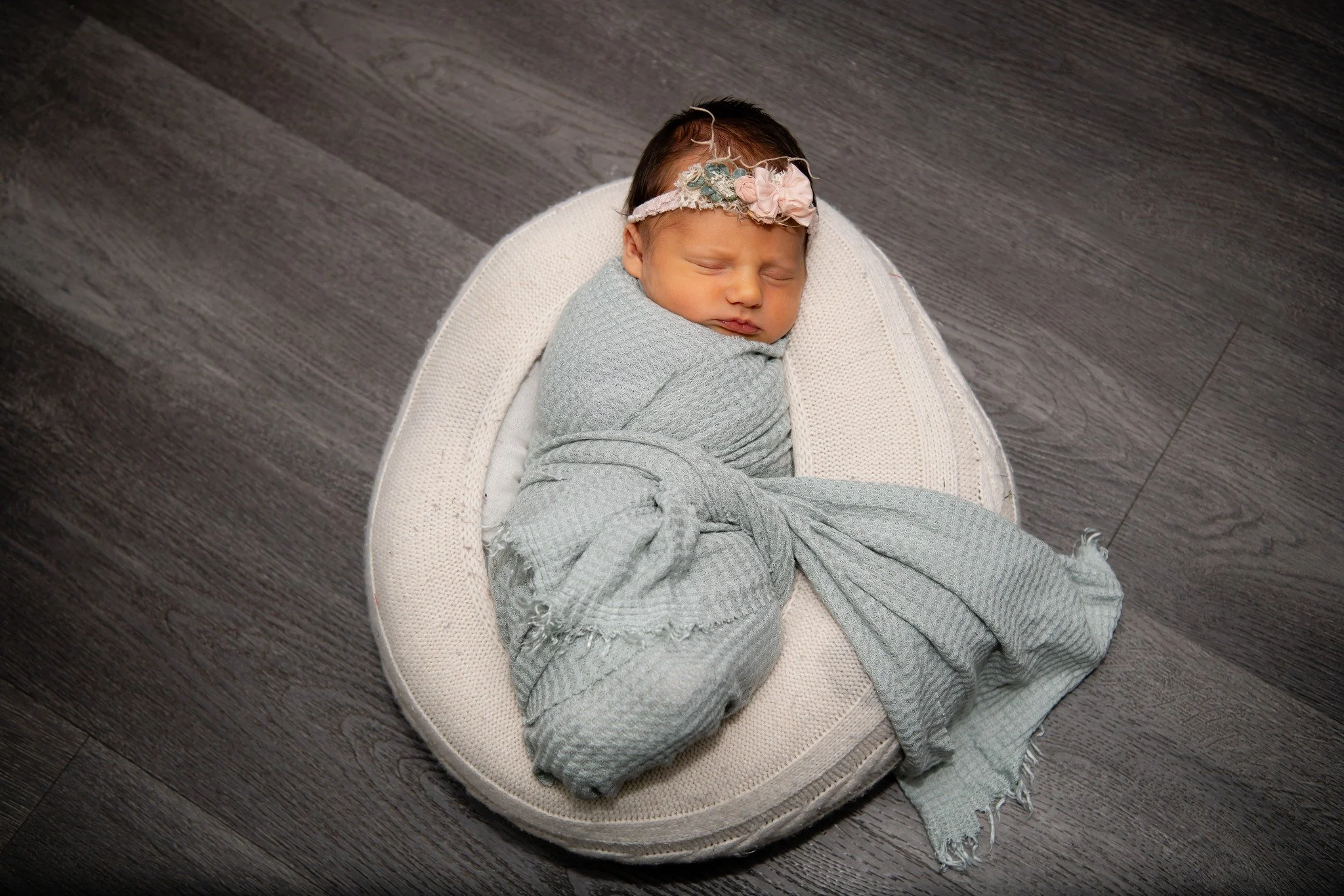 A sleeping newborn wrapped in a light gray blanket, lying on a soft, oval-shaped white cushion on a dark wooden floor. The baby is wearing a floral headband with pink and soft green accents.