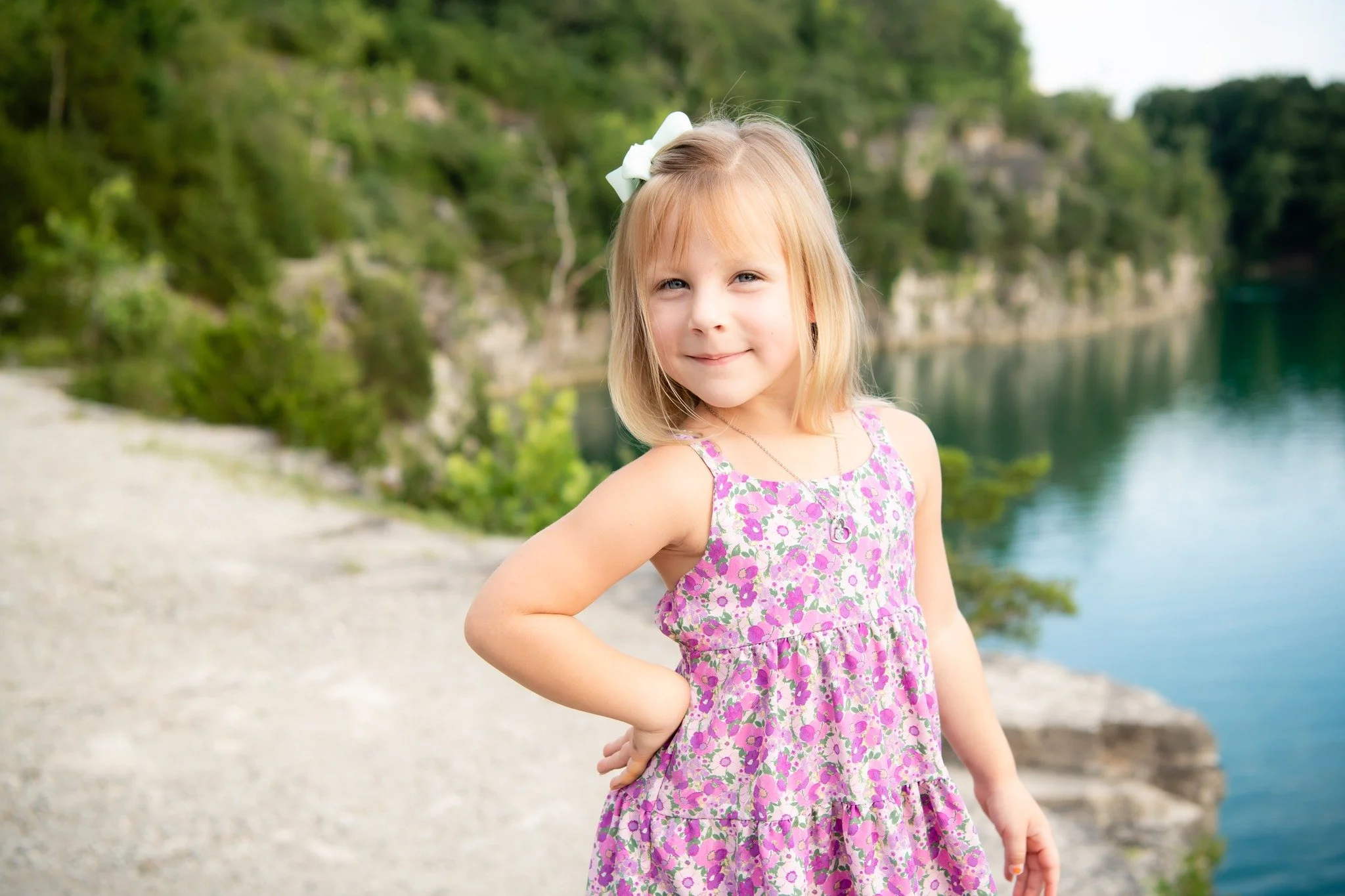 A young girl with blonde hair wearing a pink floral dress and a white bow in her hair, standing outdoors near a body of water with trees and rocky cliffs in the background.