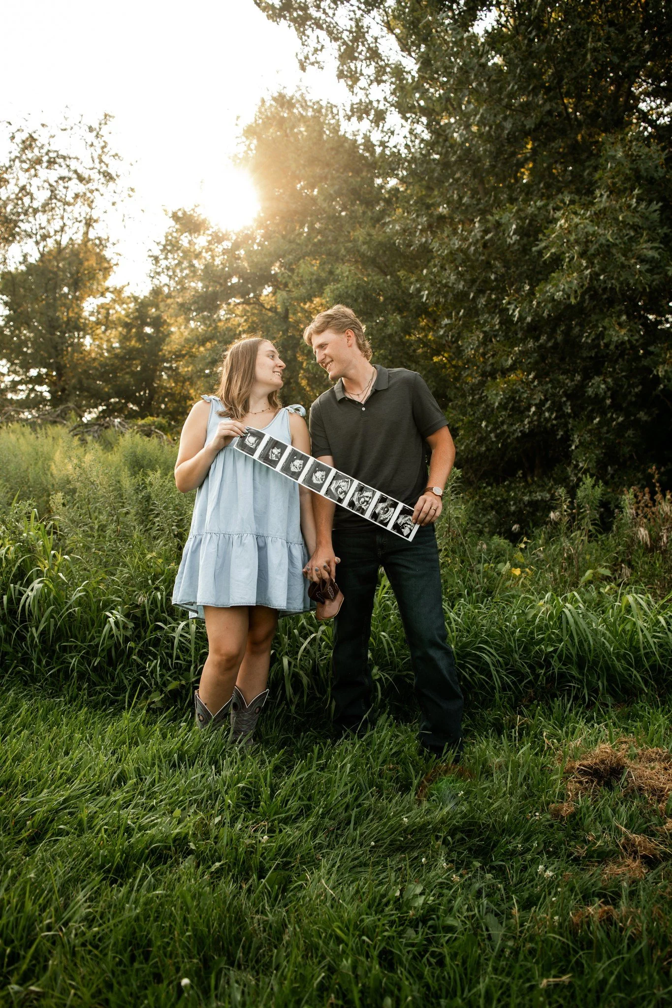 A young couple stands outdoors in a grassy field during sunset, holding ultrasound images and a sonogram, smiling at each other.
