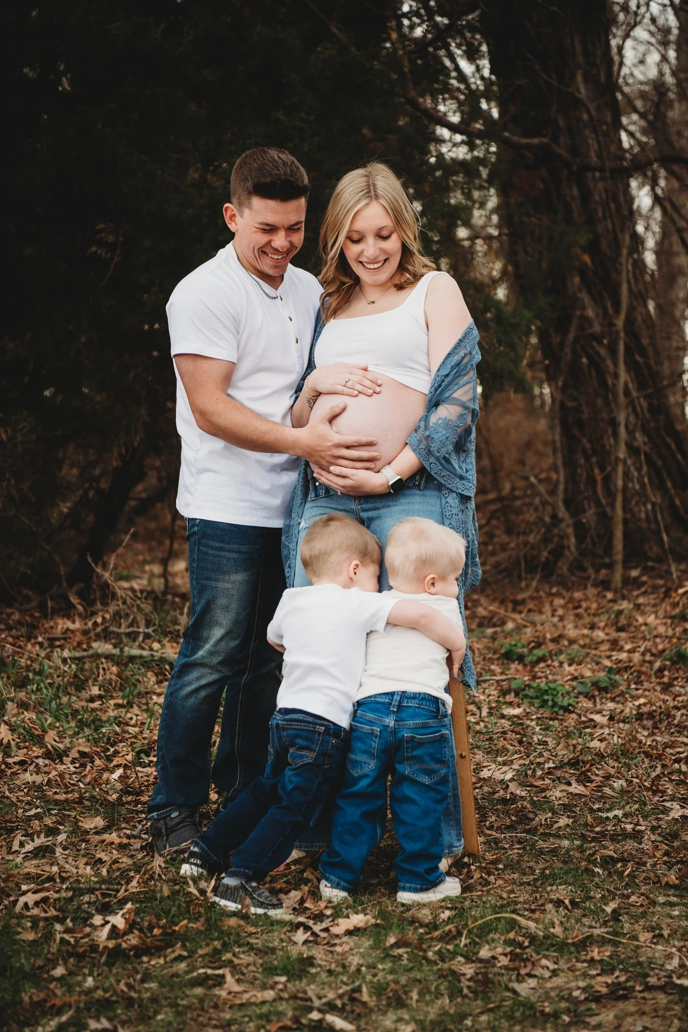 A happy family with a pregnant woman, her partner, and two young children outdoors in a wooded area during fall.