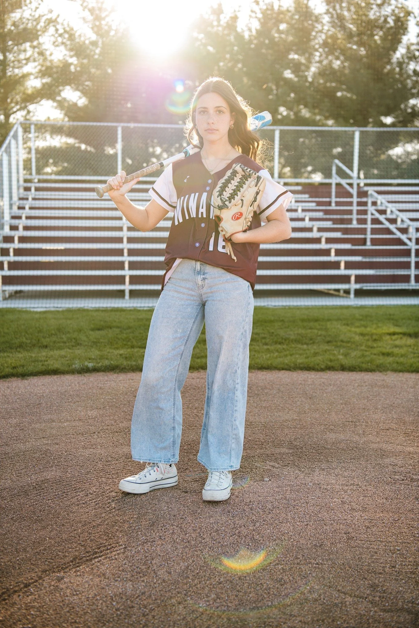 Young woman in baseball uniform holding a bat and glove on a baseball field with bleachers in the background and the sun setting behind her.