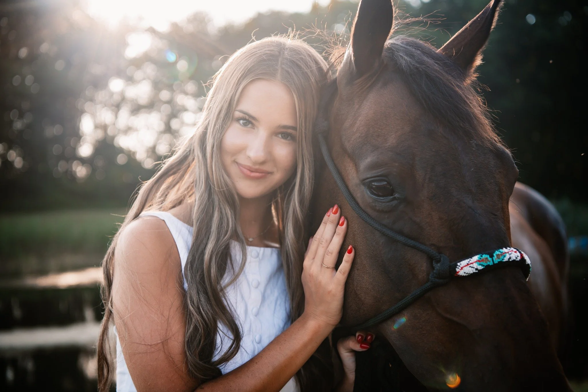 A woman with long wavy hair and red nail polish, wearing a white sleeveless top, standing next to a dark brown horse with a colorful halter. The woman is gently touching the horse's face and smiling at the camera with sunlight in the background.