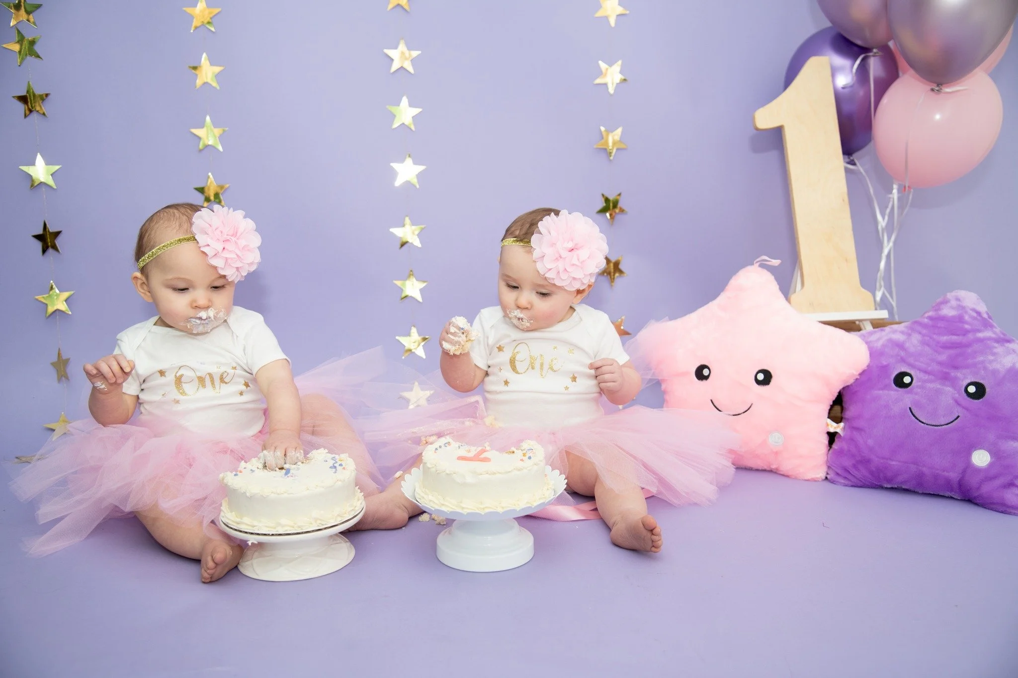 Two babies dressed in white shirts with gold writing and pink tutus celebrating their first birthday. They are sitting with cakes and surrounded by star decorations, pink and purple pillows with smiling faces, and a large wooden number one. Balloons 