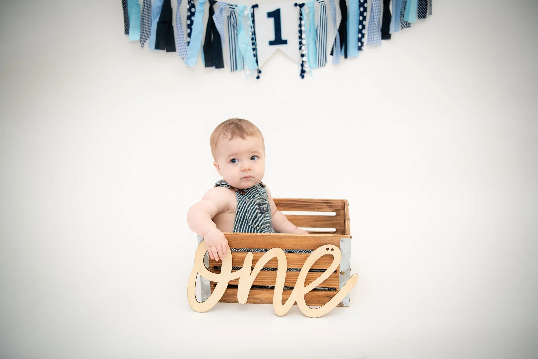 A baby sitting in a wooden crate with a wooden 'one' sign, in front of a plain white background with blue and black fabric decorations hanging above.