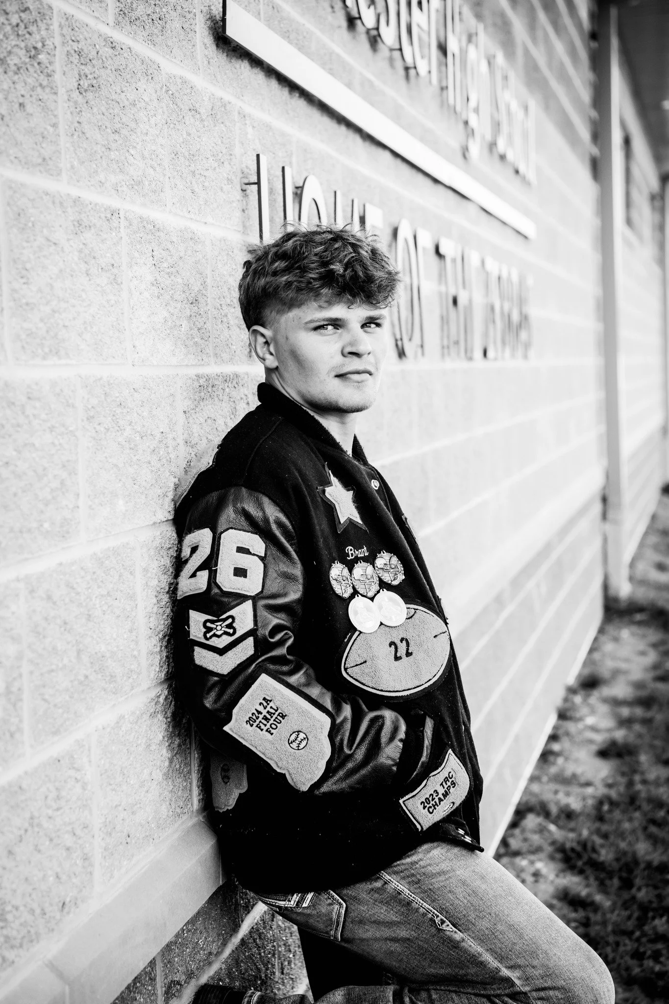 Black and white photo of a young man leaning against a brick wall, looking at the camera. He is wearing a varsity jacket with various patches and medals.